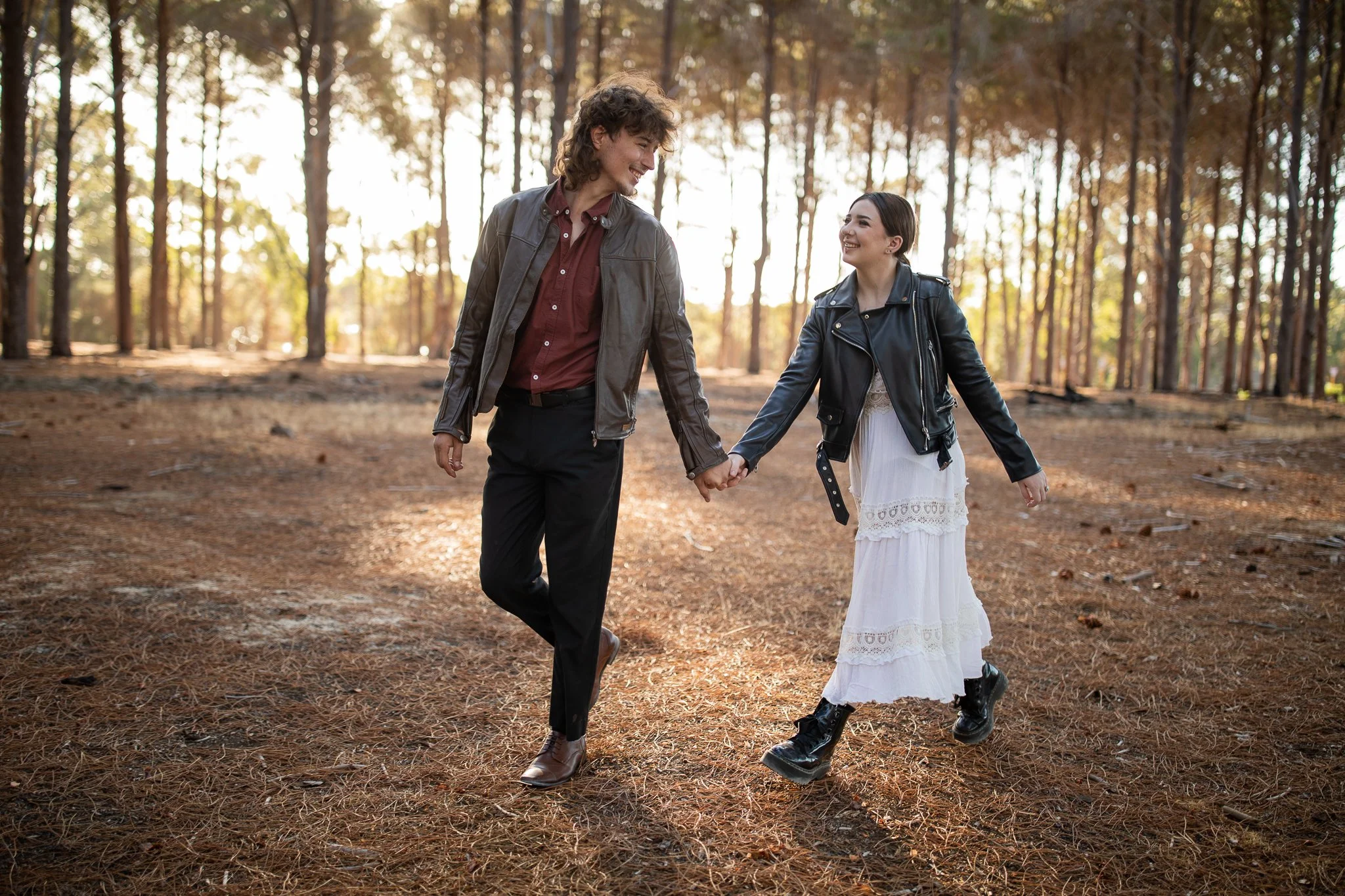 A couple holding hands and walking through a forest during fall, both wearing leather jackets and smiling at each other.