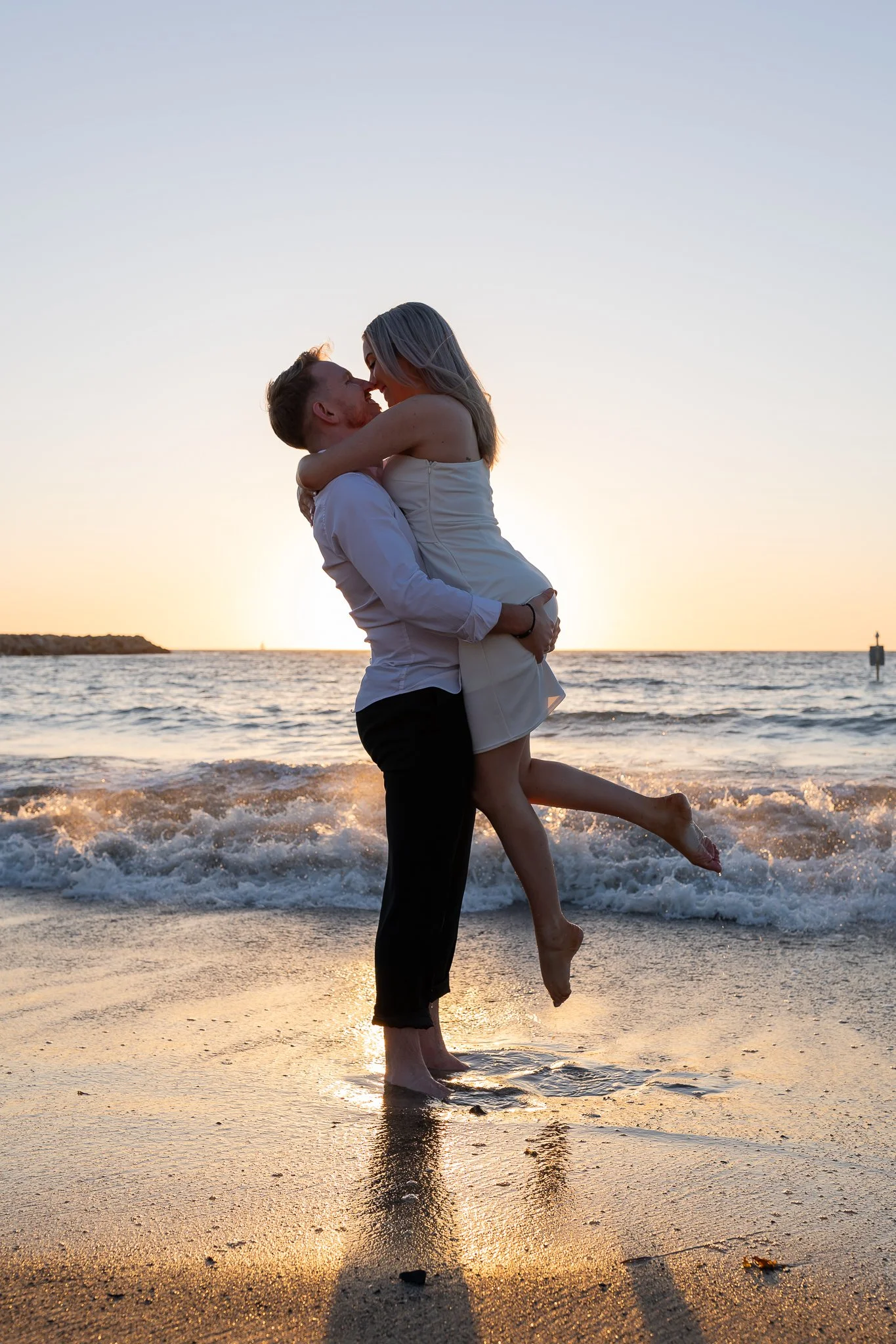 A couple embracing on the beach during sunset, with the man lifting the woman off the ground as they kiss, waves in the background.