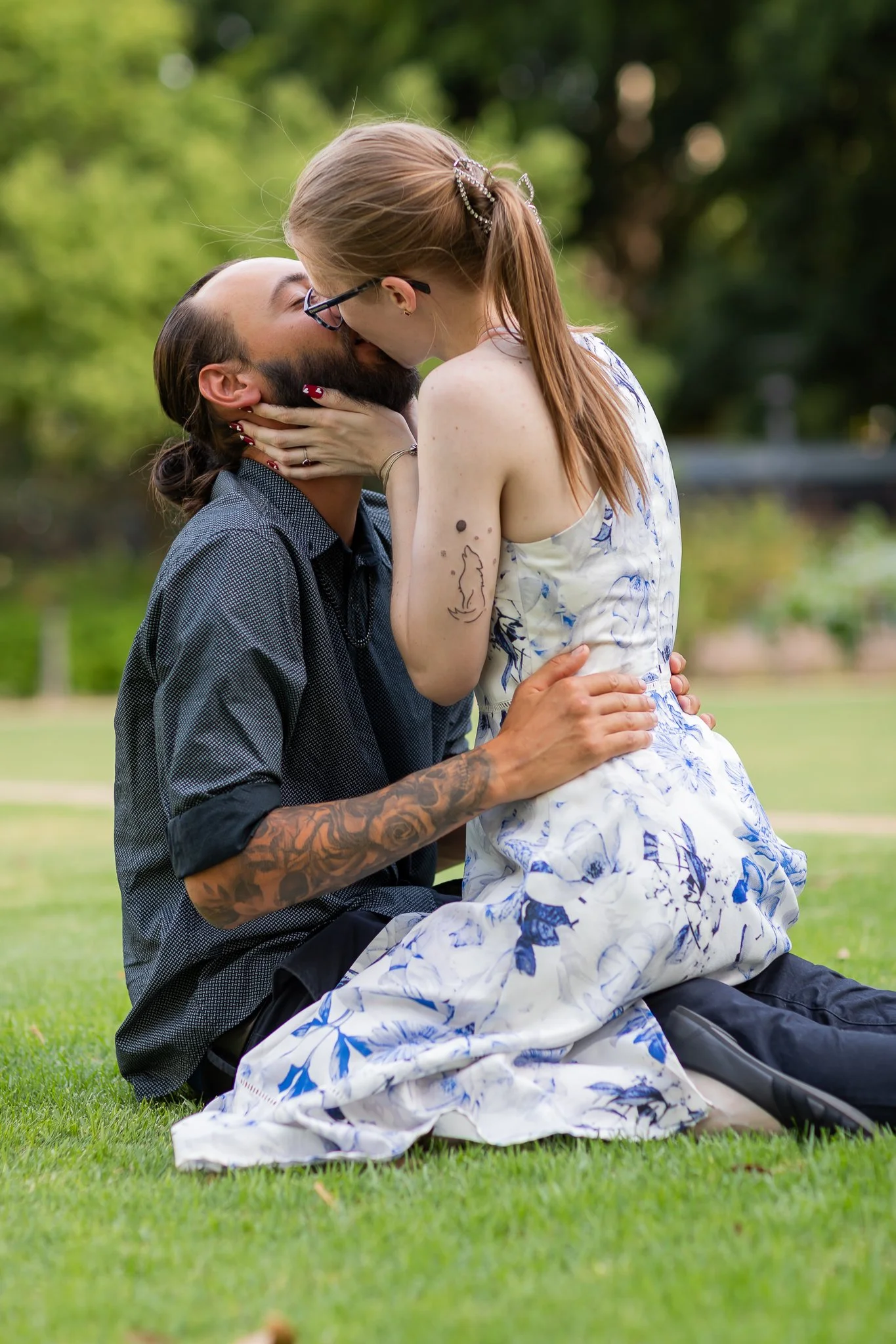 A couple sharing a kiss outdoors on the grass, with greenery in the background.