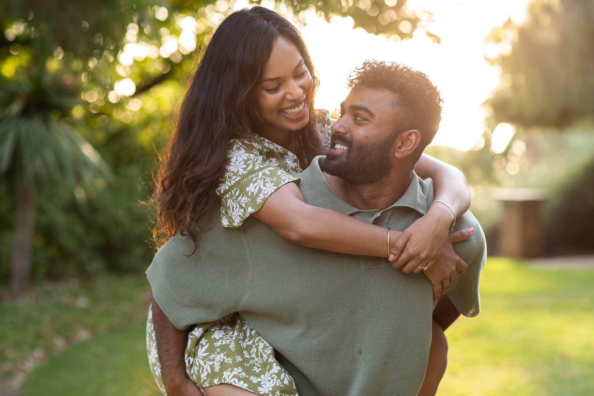 A couple smiling and embracing outdoors during sunset with greenery in the background.
