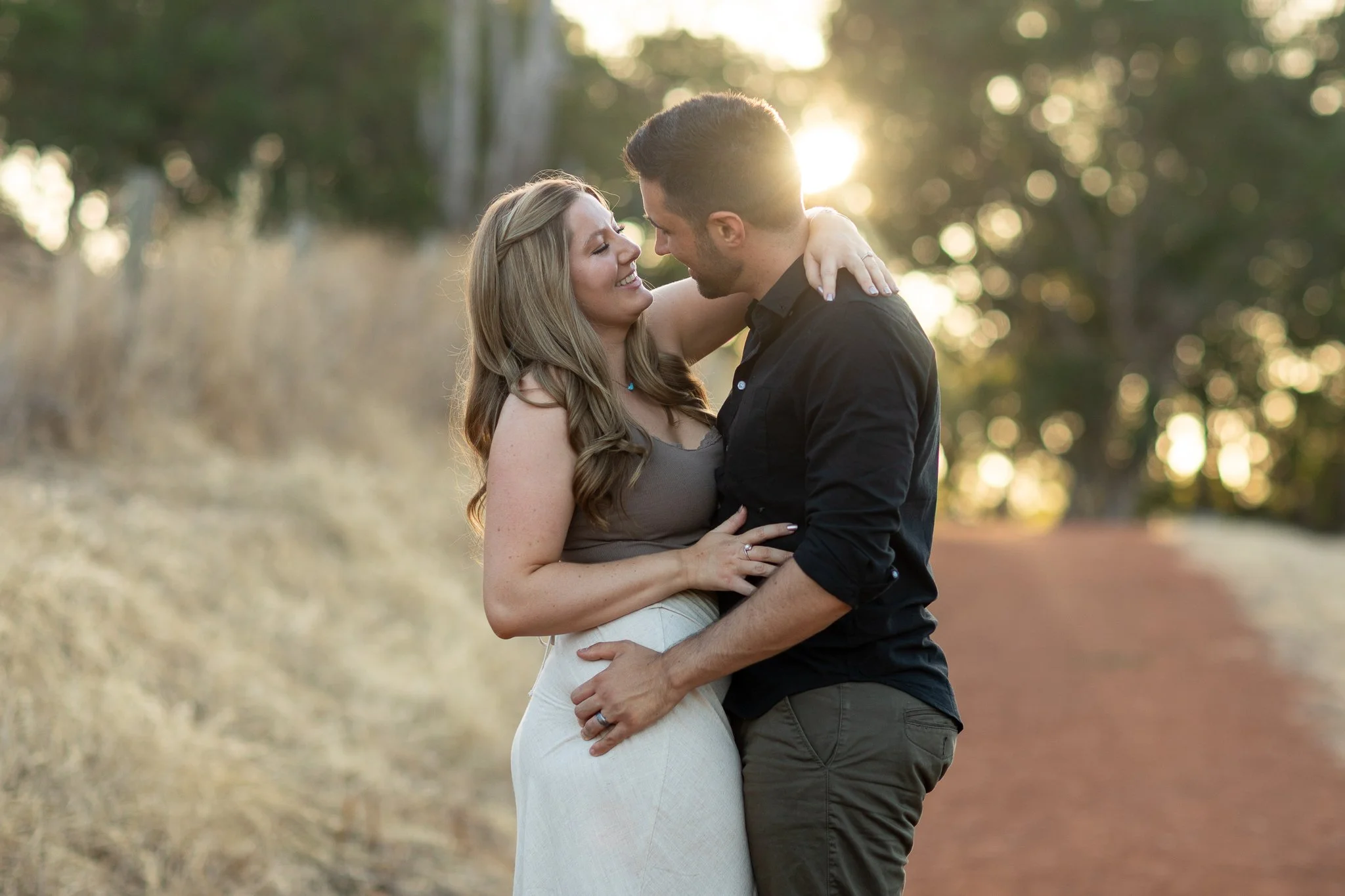 A couple embraces each other with the sun setting behind them in a wooded outdoor area.