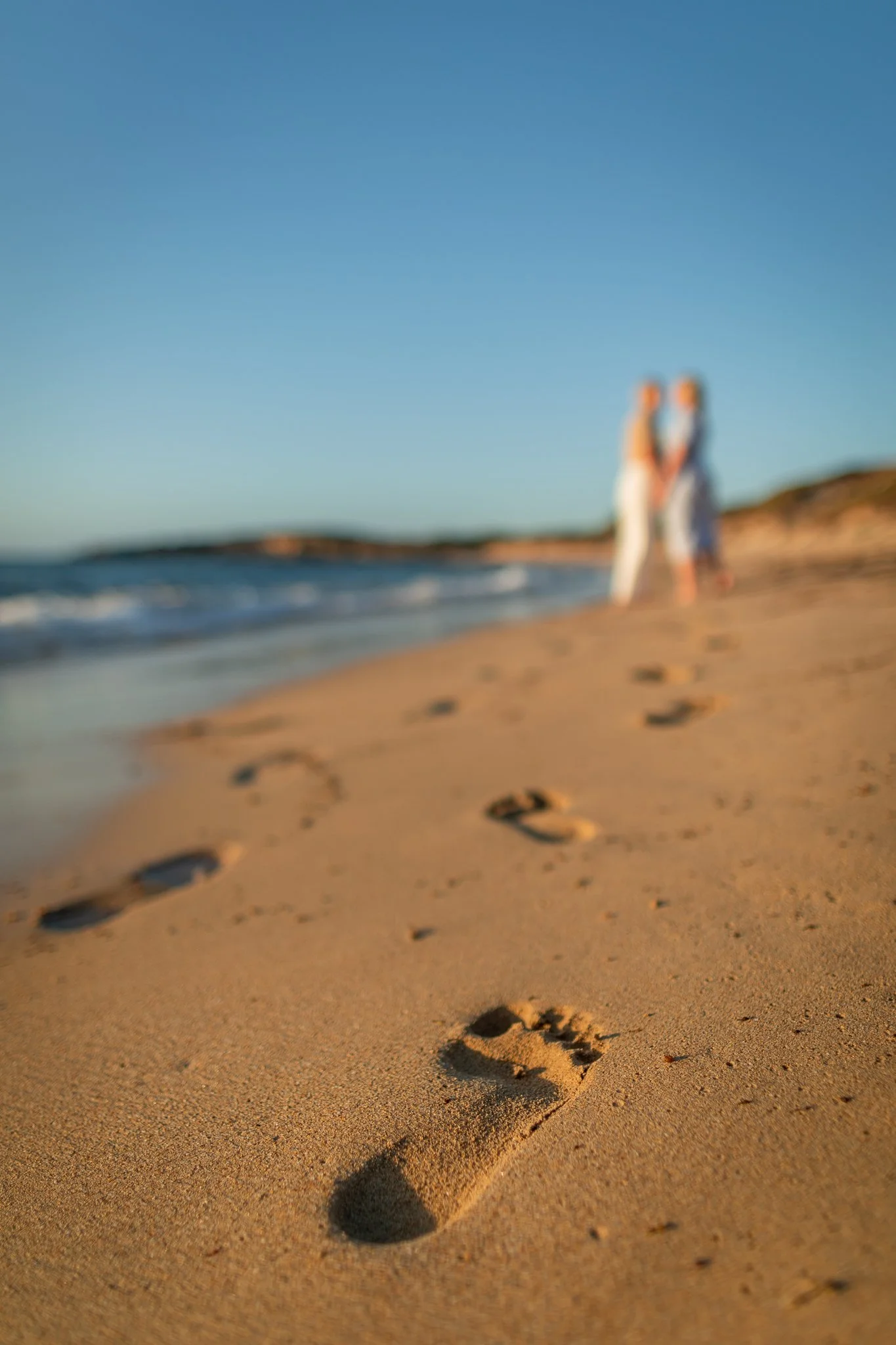 Footprints in the sand on a beach leading to two people holding hands in the distance, blurring into the background, with a clear blue sky.