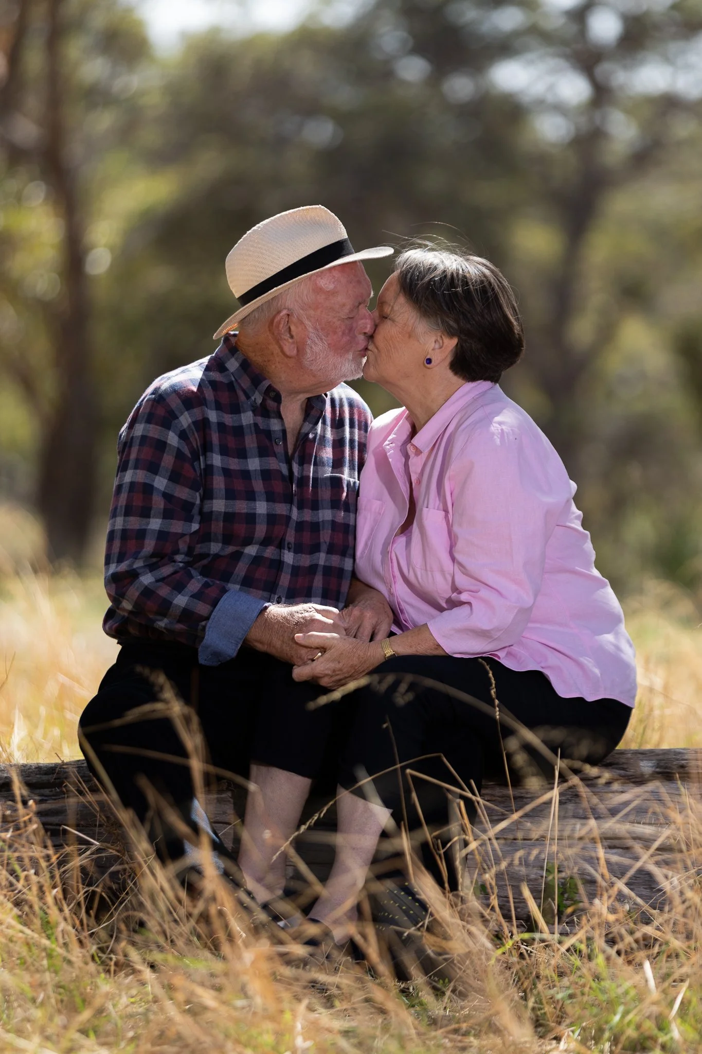 An elderly couple, sitting on a bench outdoors, sharing a kiss with trees and sunlight in the background.