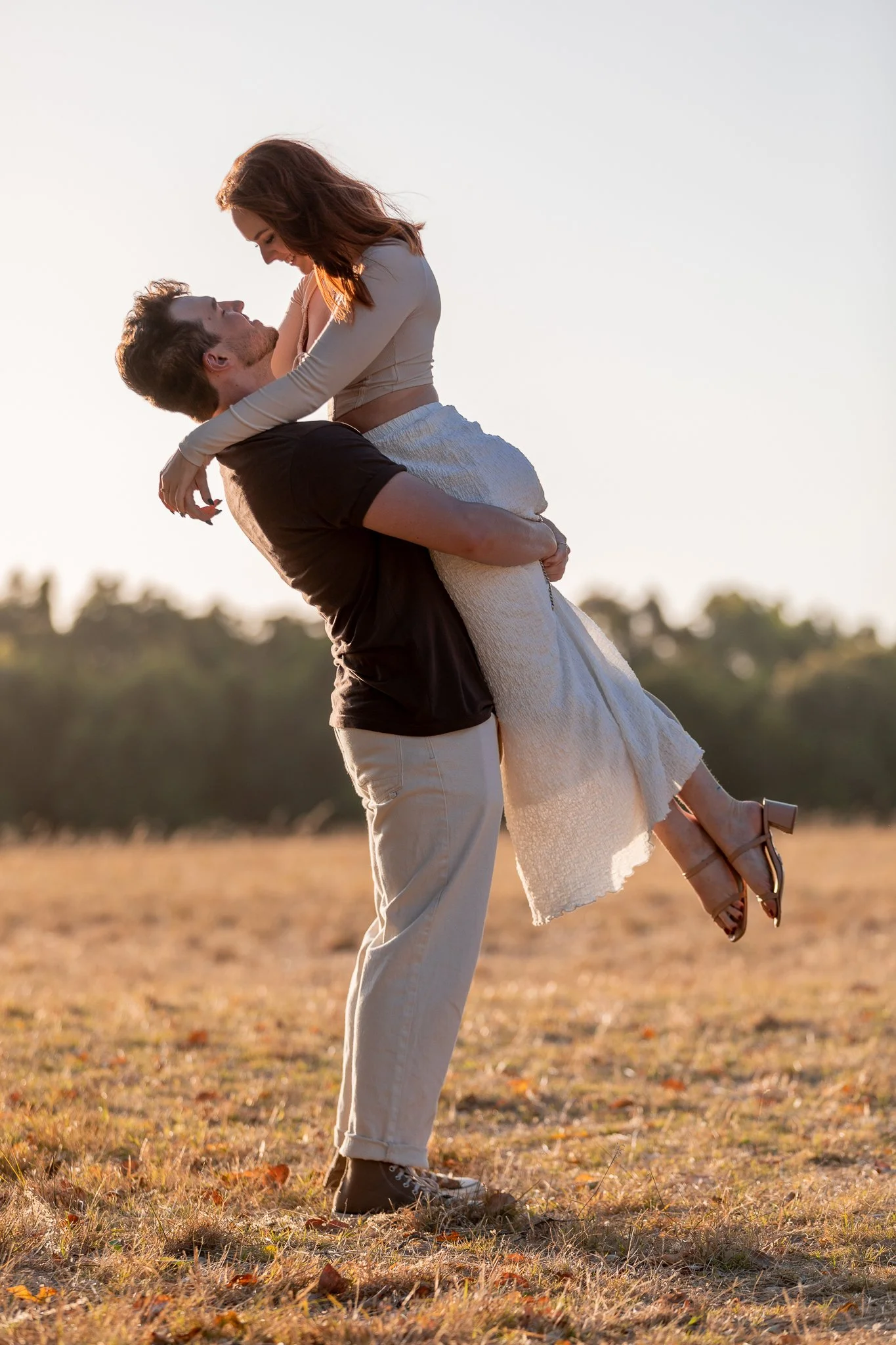 A man is holding a woman in his arms outdoors during sunset, with a blurred landscape in the background.
