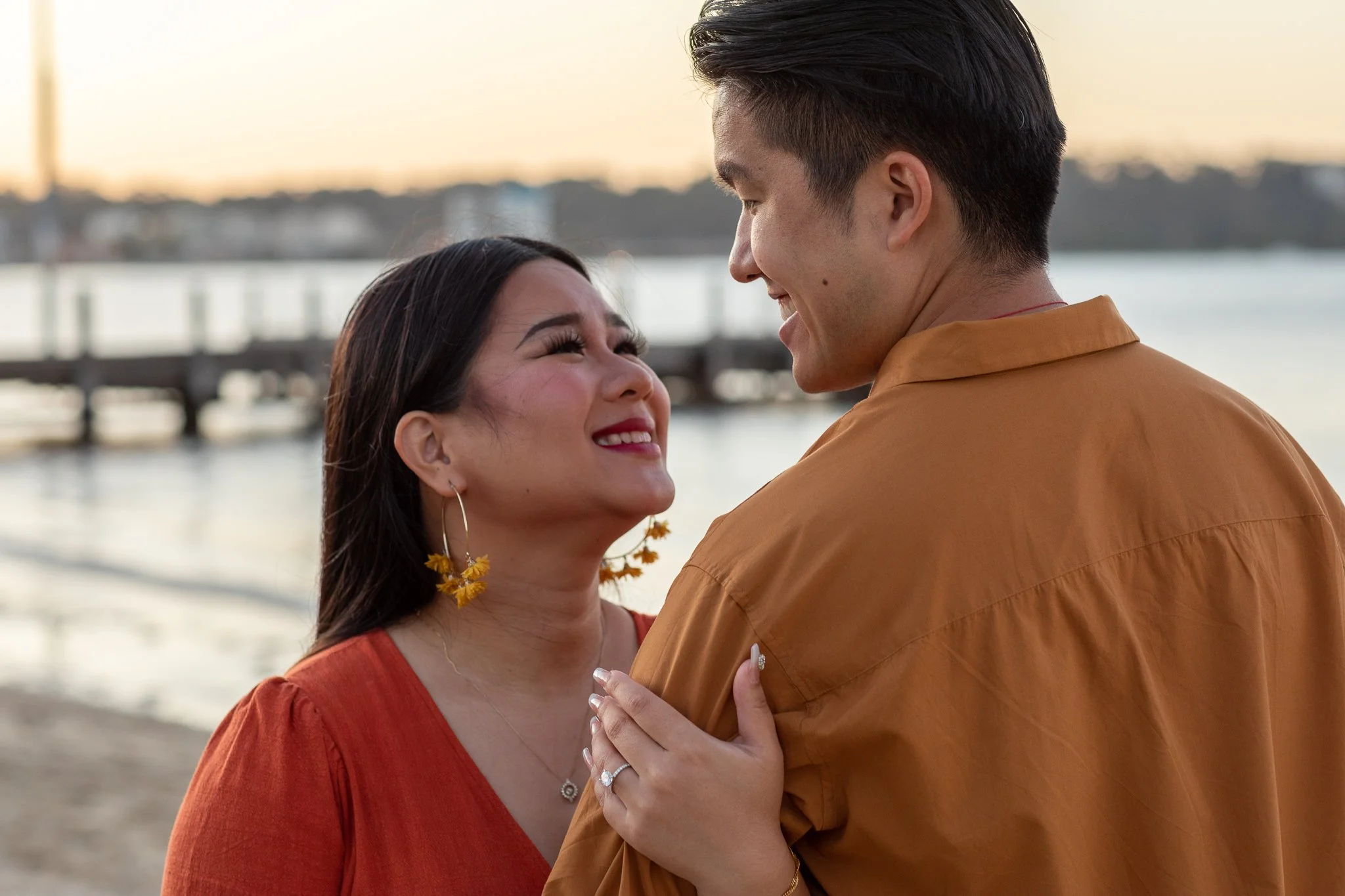 A woman and a man are looking at each other affectionately outdoors near a body of water at sunset.