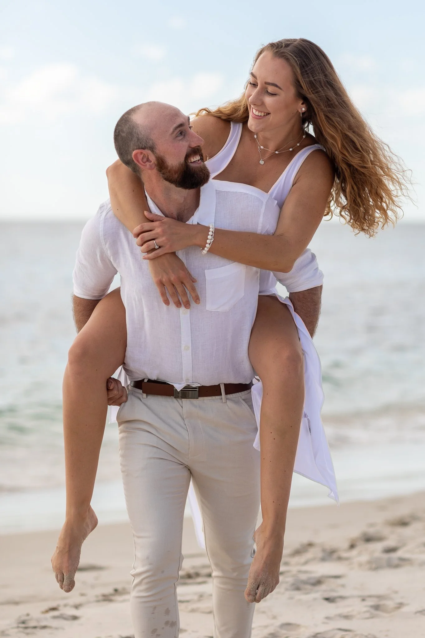 A man is carrying a woman on his back at the beach. They are smiling and looking at each other. The woman has long, wavy hair and is wearing a white dress. The man has a beard, is bald, and is wearing a white shirt and beige pants. The background sho