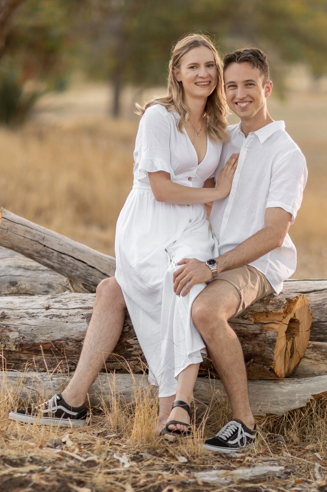 A young couple sitting on a log outdoors, smiling and looking at the camera, with a blurred background of trees and dry grass.