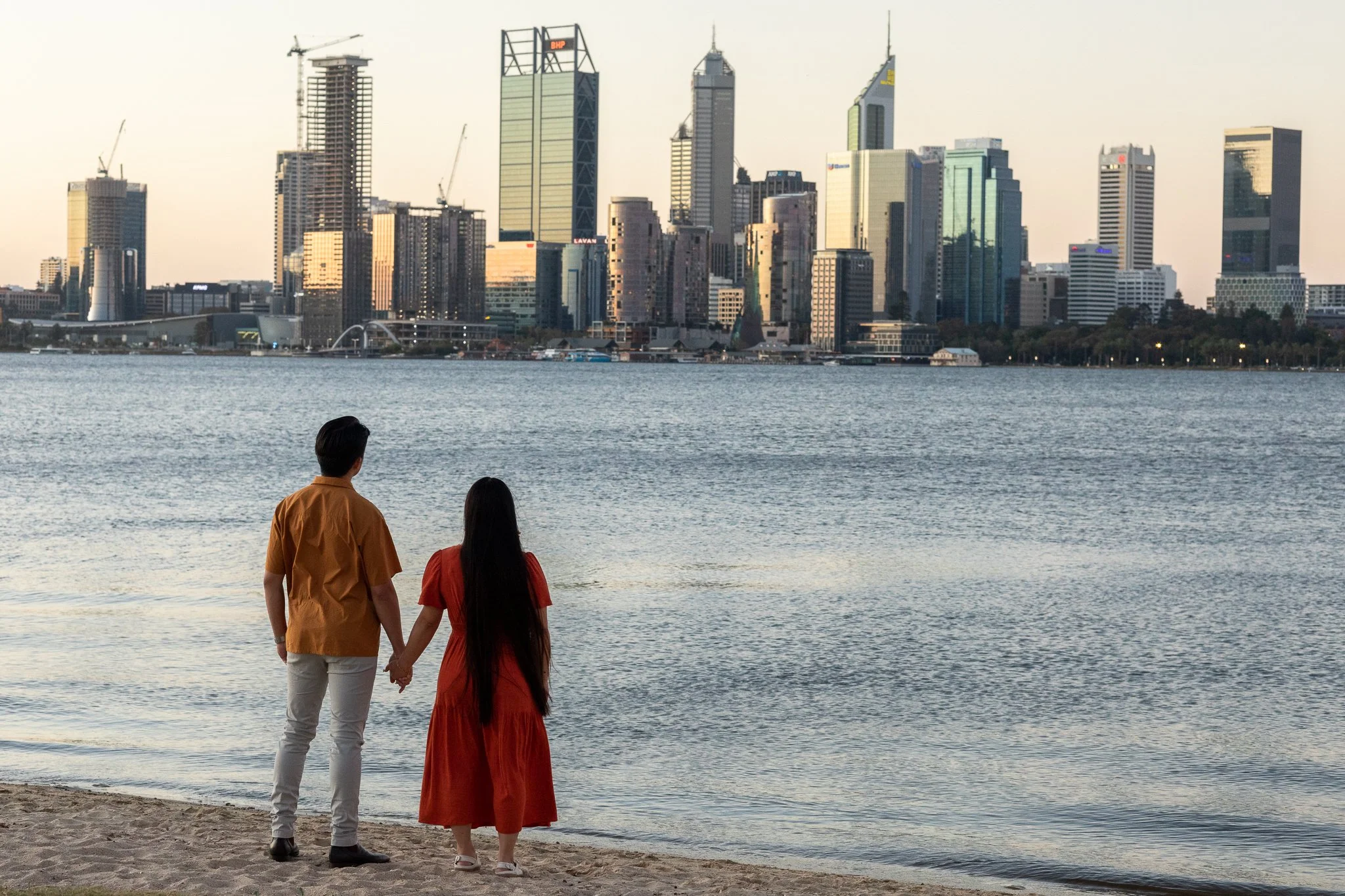 A couple holding hands on the beach, overlooking a city skyline across the water