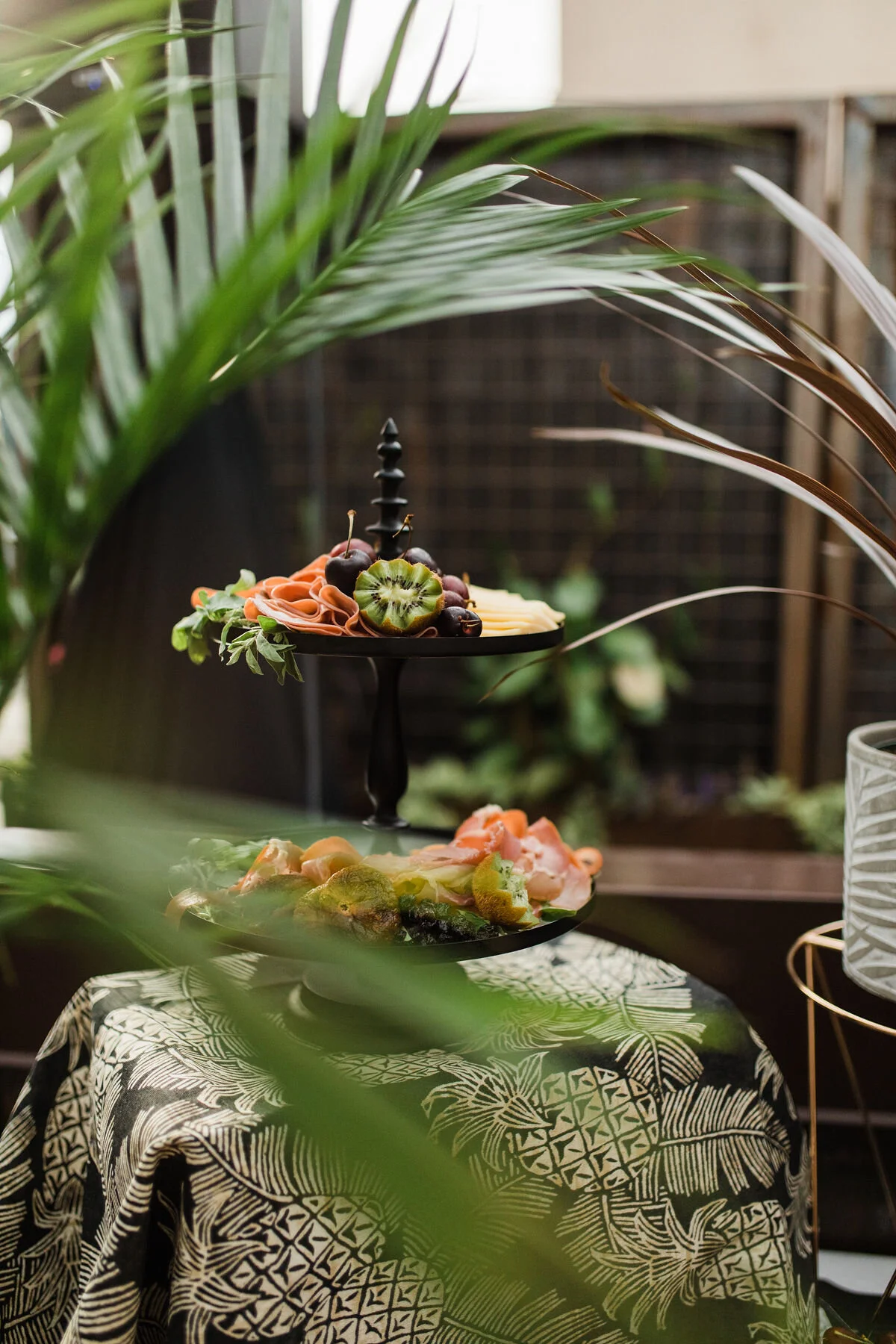 Two-tiered black serving stand with assorted sliced fruits, including kiwi, cherries, and grapes, placed on a table with a black and white pineapple-patterned tablecloth, surrounded by green tropical plants in an outdoor setting.