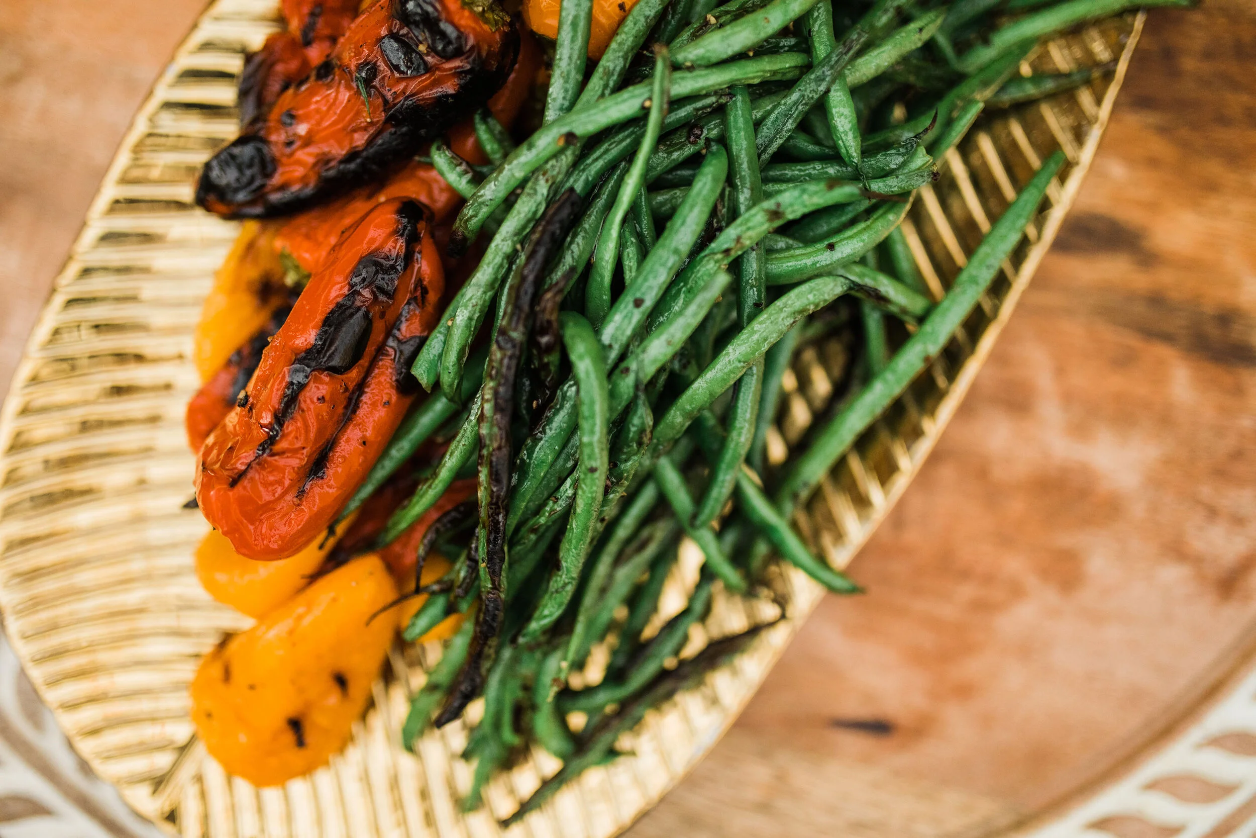Grilled red, yellow, and orange peppers and green beans on a woven wooden platter.