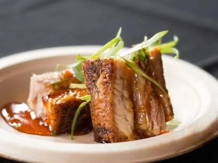 A plate with a slice of cooked pork belly topped with green garnishes and drizzled with sauce.
