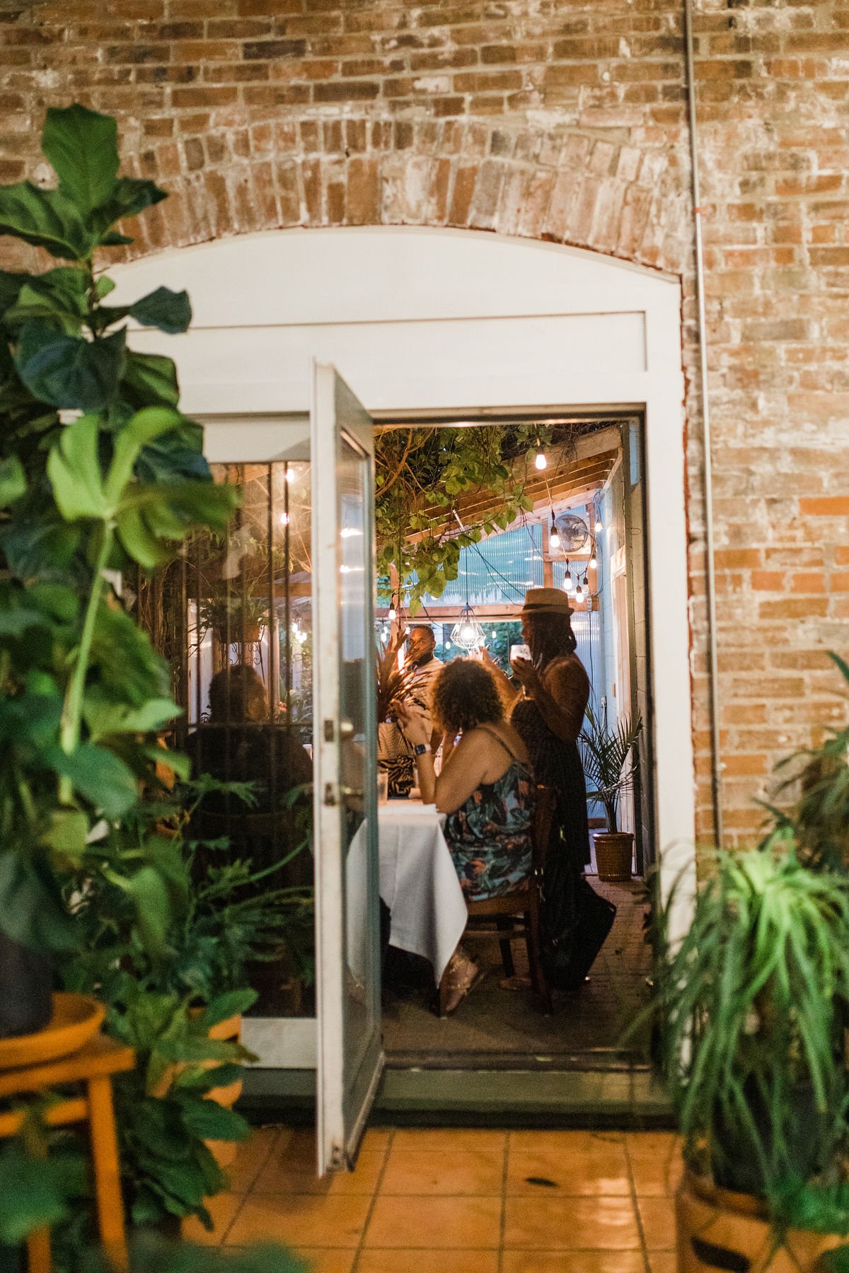 People dining inside a cozy restaurant through an open glass door, with indoor plants and warm string lights creating an inviting atmosphere.