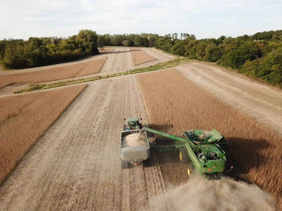 Aerial view of a green combine harvester working in a field during harvest with a truck waiting to unload grain, surrounded by farmland and trees.