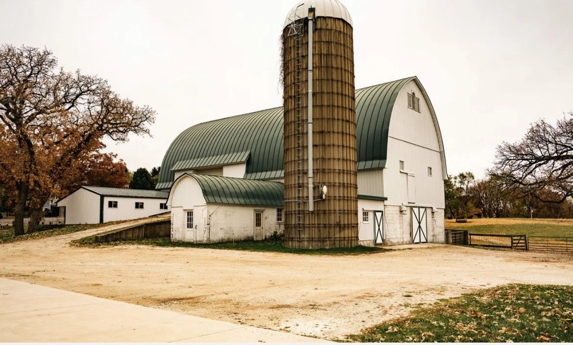 A white barn with green roofing, a tall silo, leafless trees, and a dirt ground under an overcast sky.