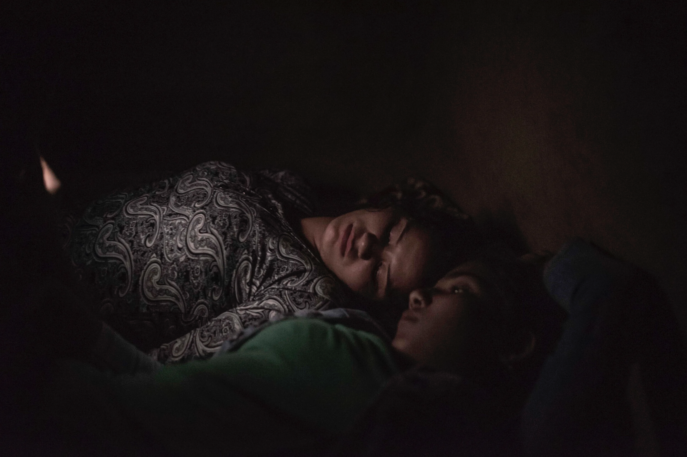 Two children lying close together, one with glasses, in a dark setting, resting or sleeping.
