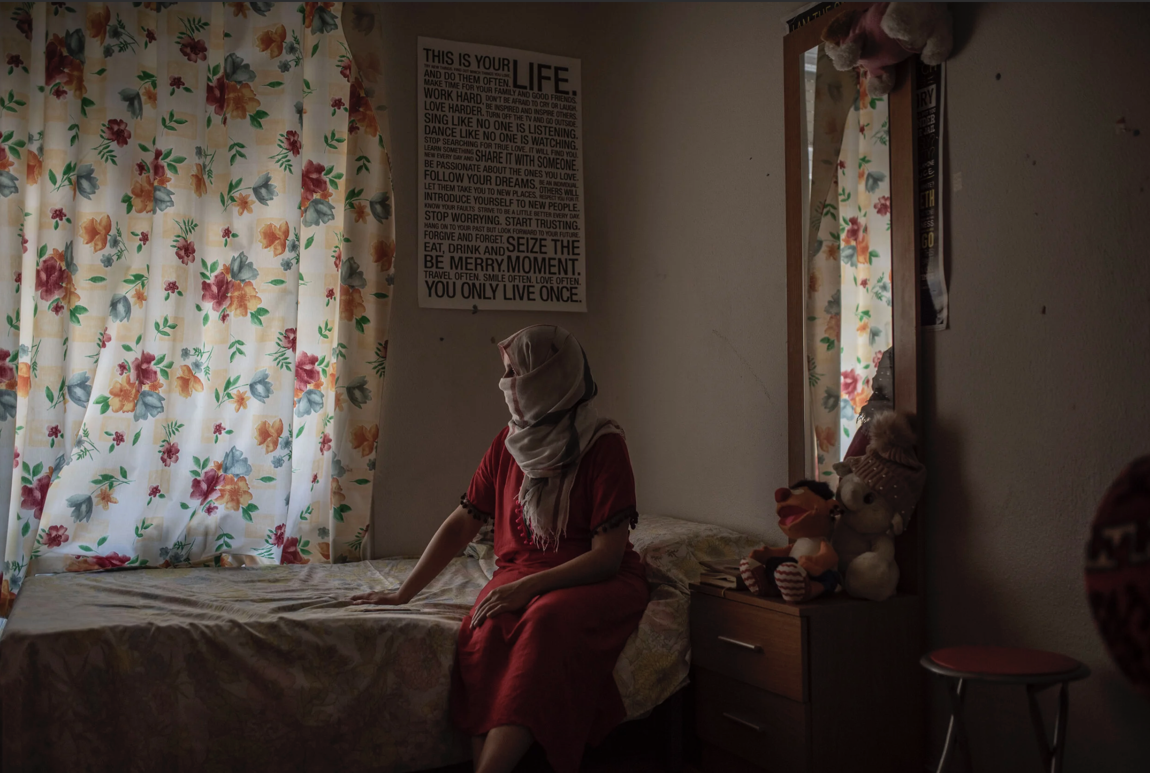 A woman sitting on a bed in a bedroom with floral curtains, a poster on the wall with motivational quotes, and stuffed animals on a nightstand.