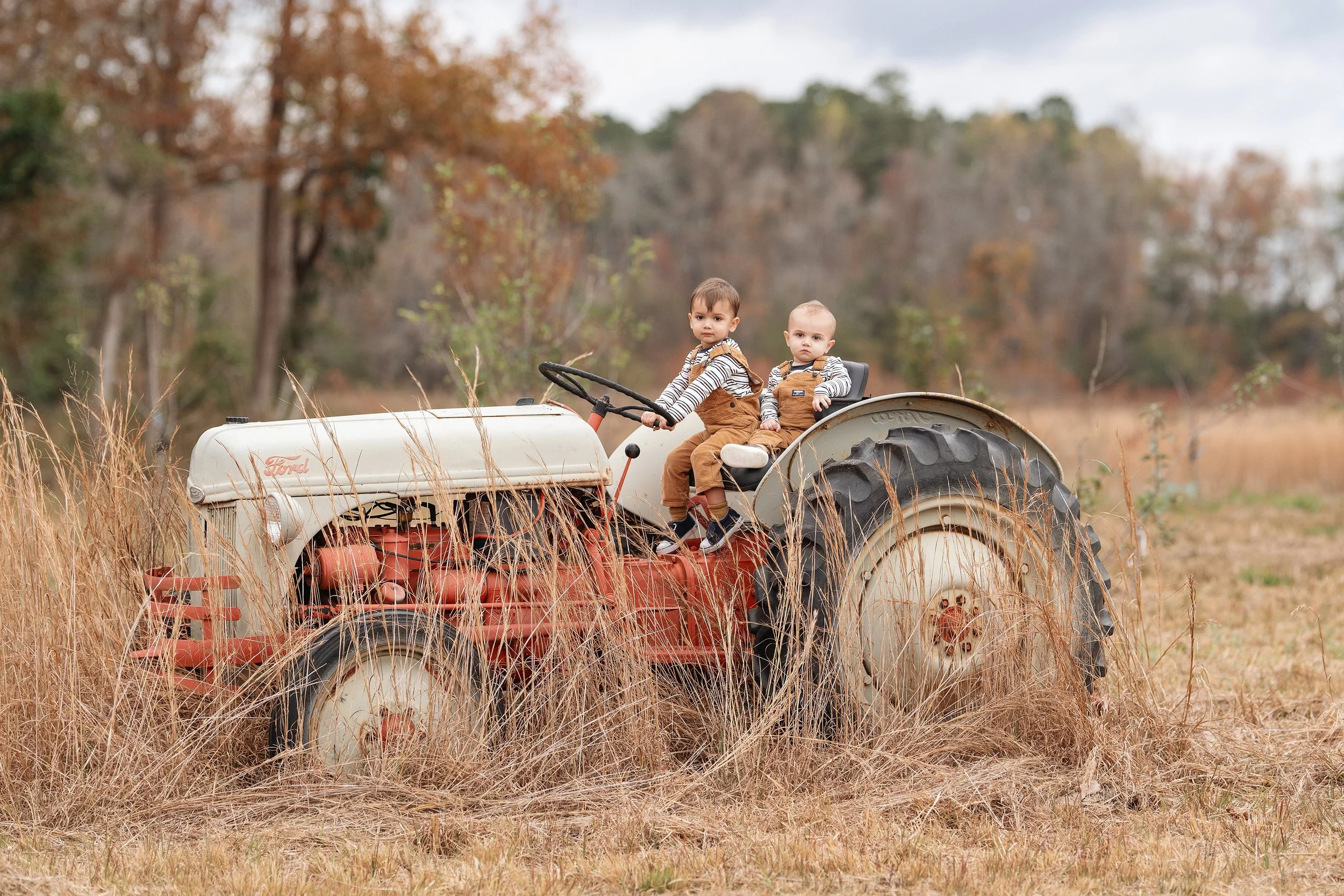 rustic outdoor portrait of two young siblings sitting on a vintage tractor in a grassy field. Dressed in matching striped tops and corduroy overalls, this playful children's photosession was captured by Sommer Ortiz Portraits in North Myrtle Beach, S