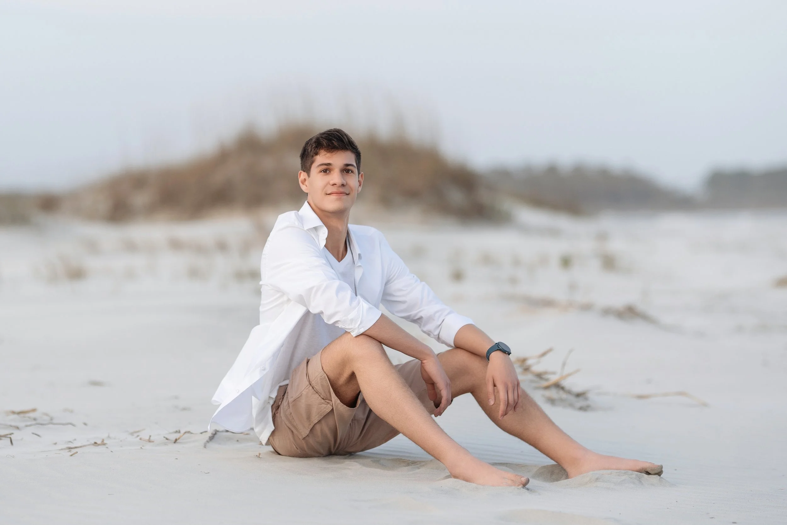 Relaxed and joyful highschool senior sits on the sun-kissed sands of Myrtle Beach, dressed in a casual white shirt and khaki shorts, exuding the carefree spirit of youth. This natural outdoor setting highlights the milestone of senior year, making it