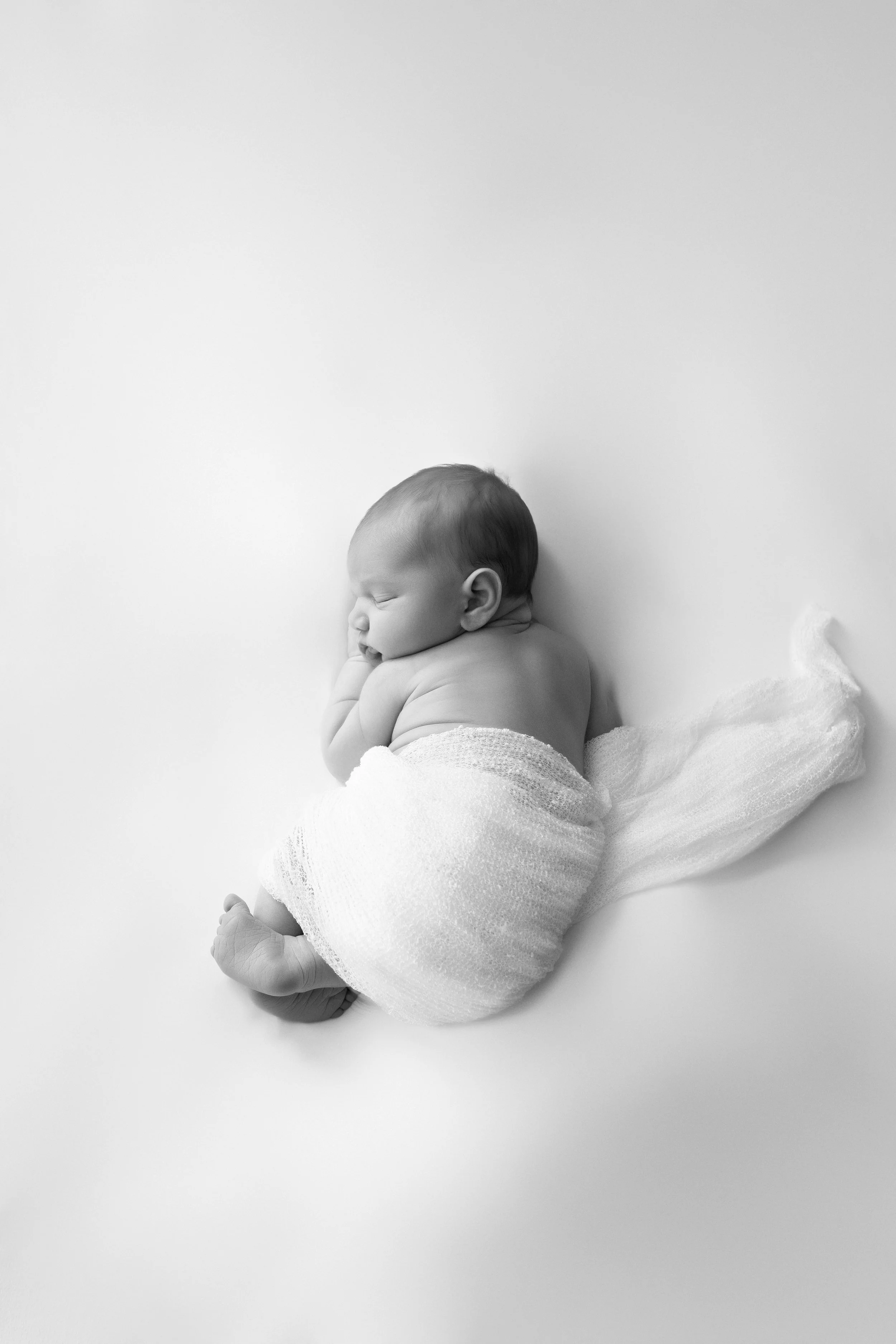 Artistic black and white image of a peaceful newborn swaddled in a delicate white cloth, curled in a natural pose that captures the essence of new life. This minimalist portrait highlights the baby's serene expression, perfect for birth announcements
