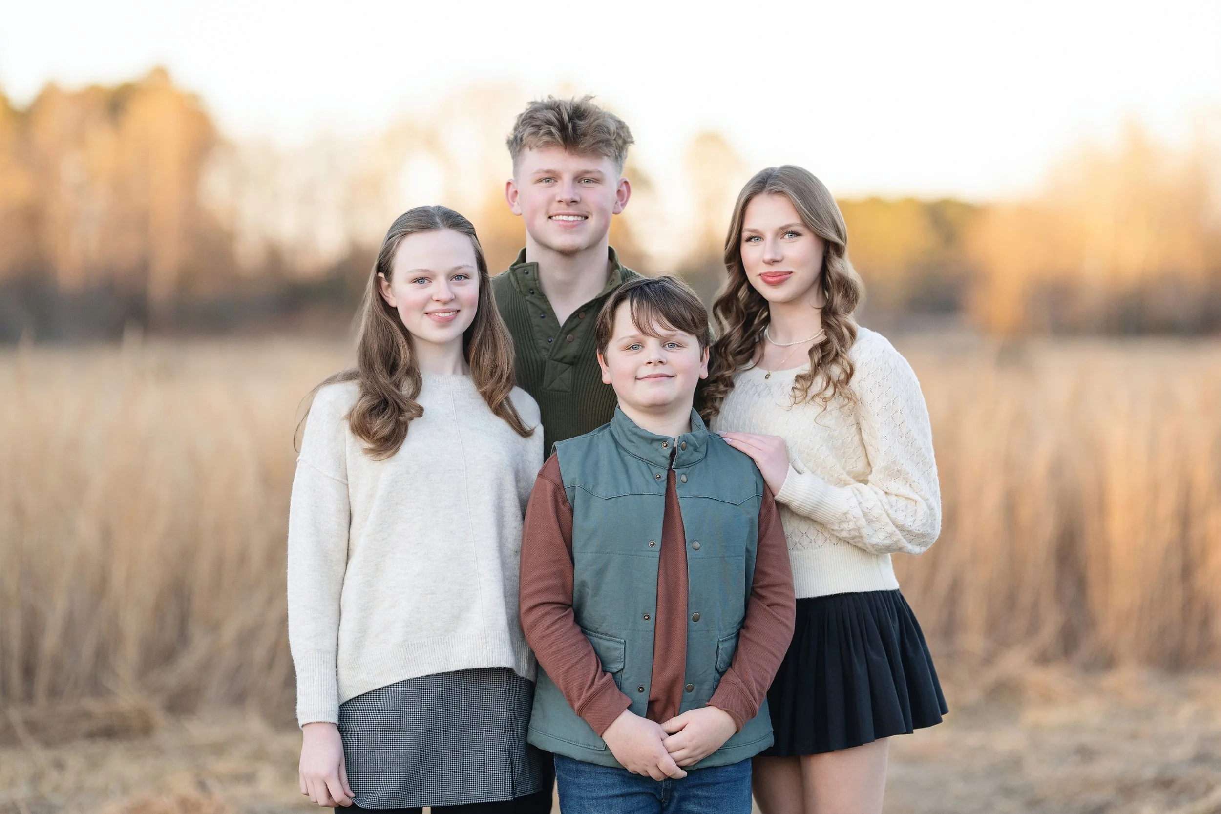 Golden hour outdoor family portrait of four siblings in coordinating fall outfits, photographed in a natural field setting near North Myrtle Beach, SC. captured by Sommer Ortiz Portraits, specializing in lifestyle family photography with soft, natura