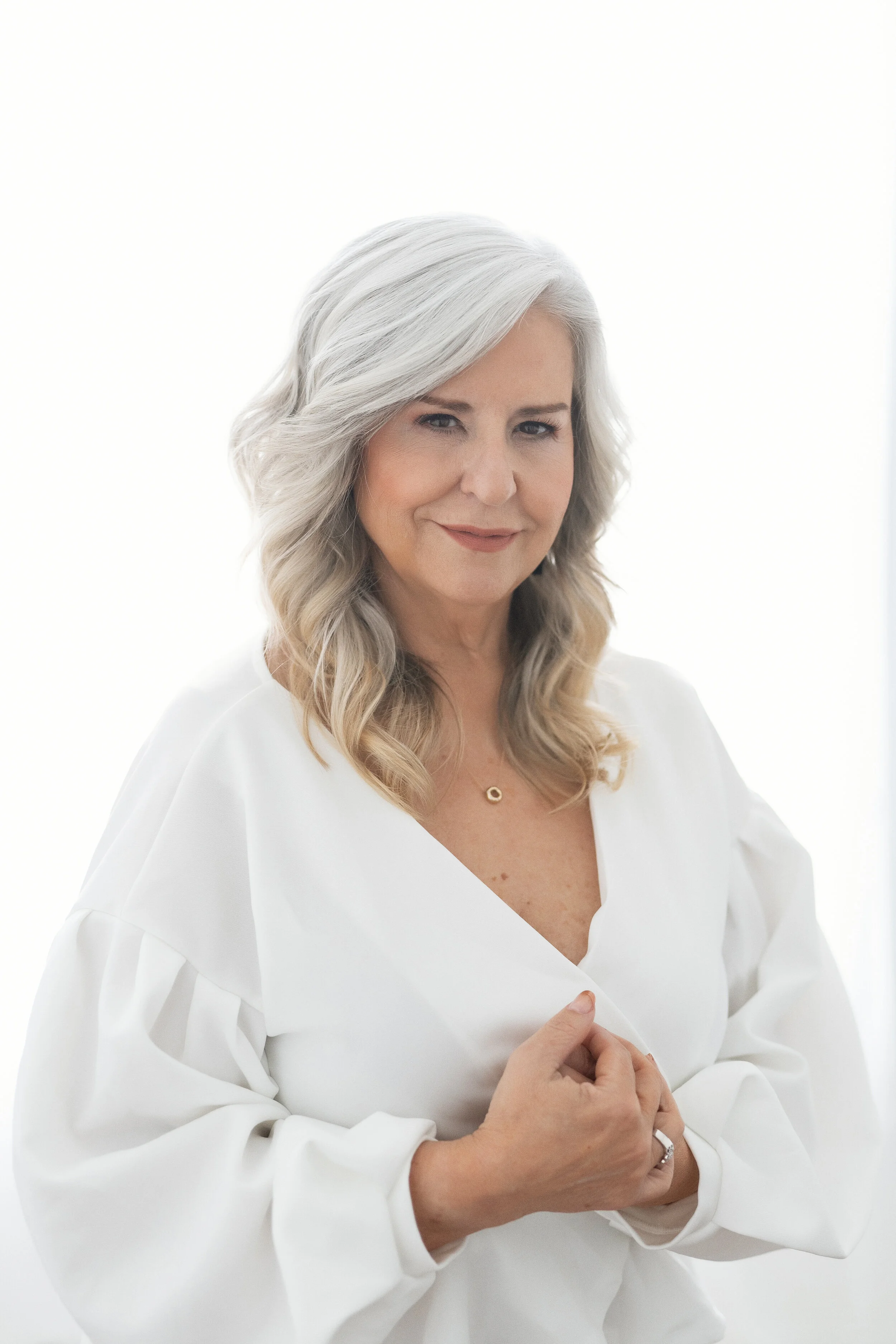 Protrait of an elegant woman with silver hair styled in soft waves, wearing a chic white blouse. The high-key lighting and her serene expression evoke a sense of confidence and grace, perfect for professional headshots or personal branding photograph