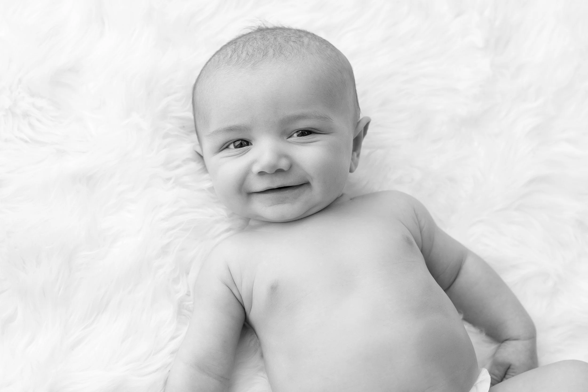 Black and white photograph showcasing the joyful smile of a baby lying on a plush white blanket. The image captures the pure and infectious happiness of infancy, ideal for a family photo album or a nursery wall portrait.