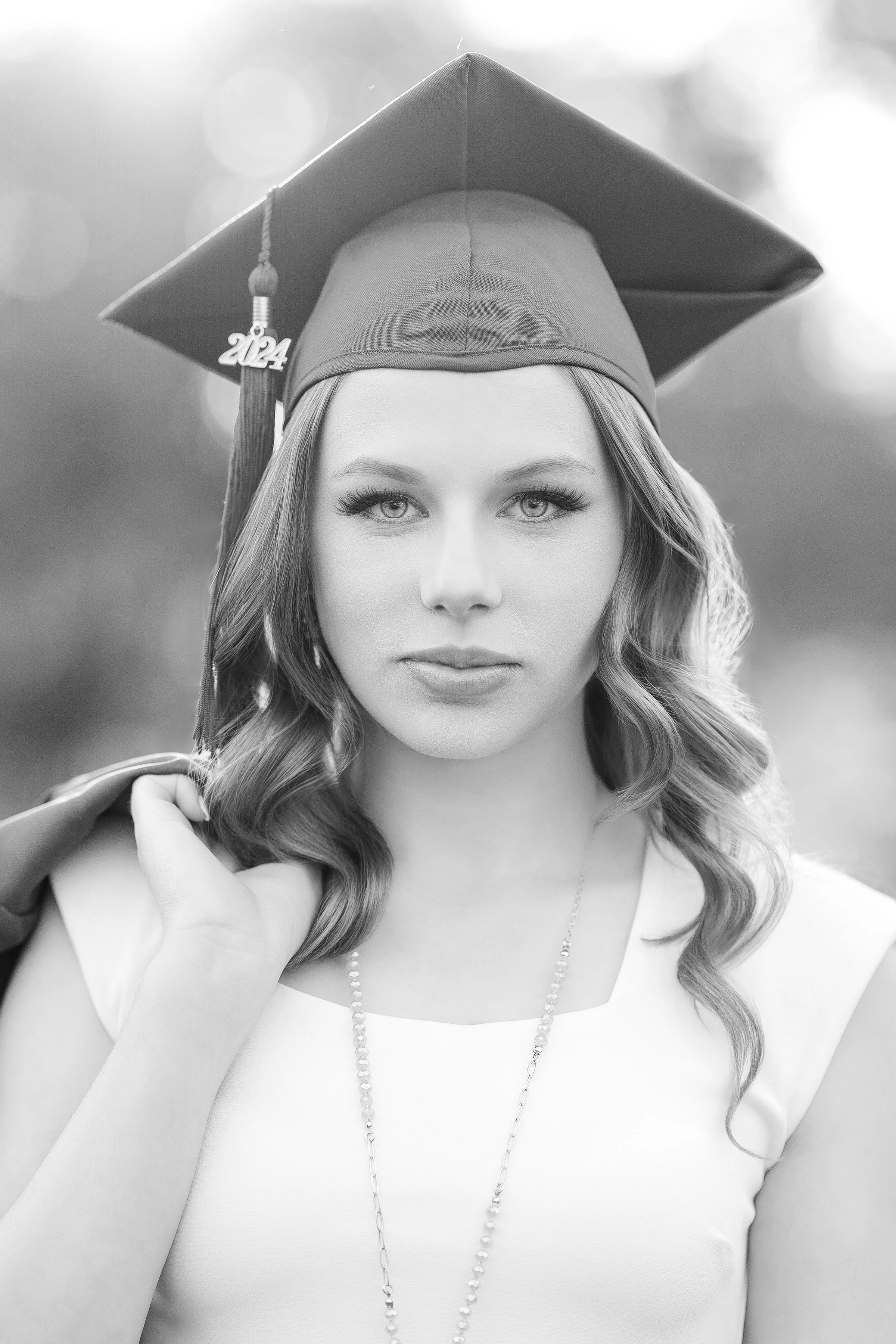 This black and white photograph captures a poised young woman in her graduation attire. She stands confidently, her gaze fixed forward, exuding a sense of accomplishment  and anticipation for the future. The academic cap with a tassel marked"2024" si