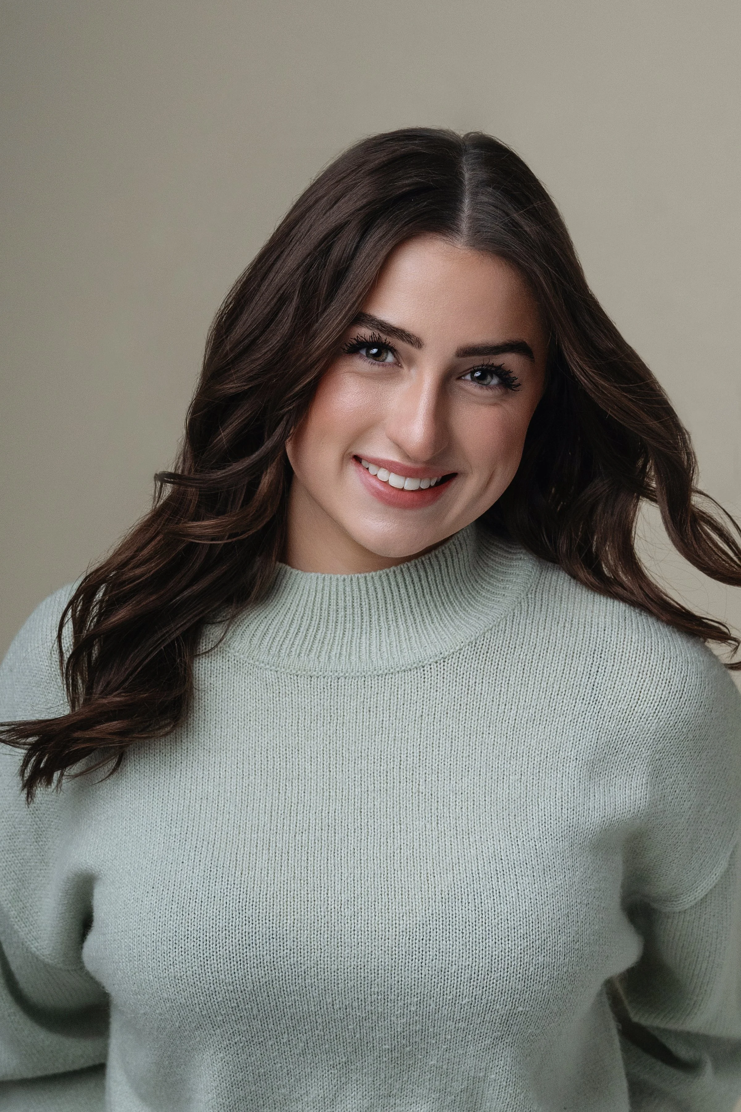 This is a professional portrait of a woman with a  vibrant smile, capturing a look thats both polished and approachable. The subject wears a soft-textured, pale green turtleneck sweater that exudes comfort and style. Her dark, wavy hair frames her fa