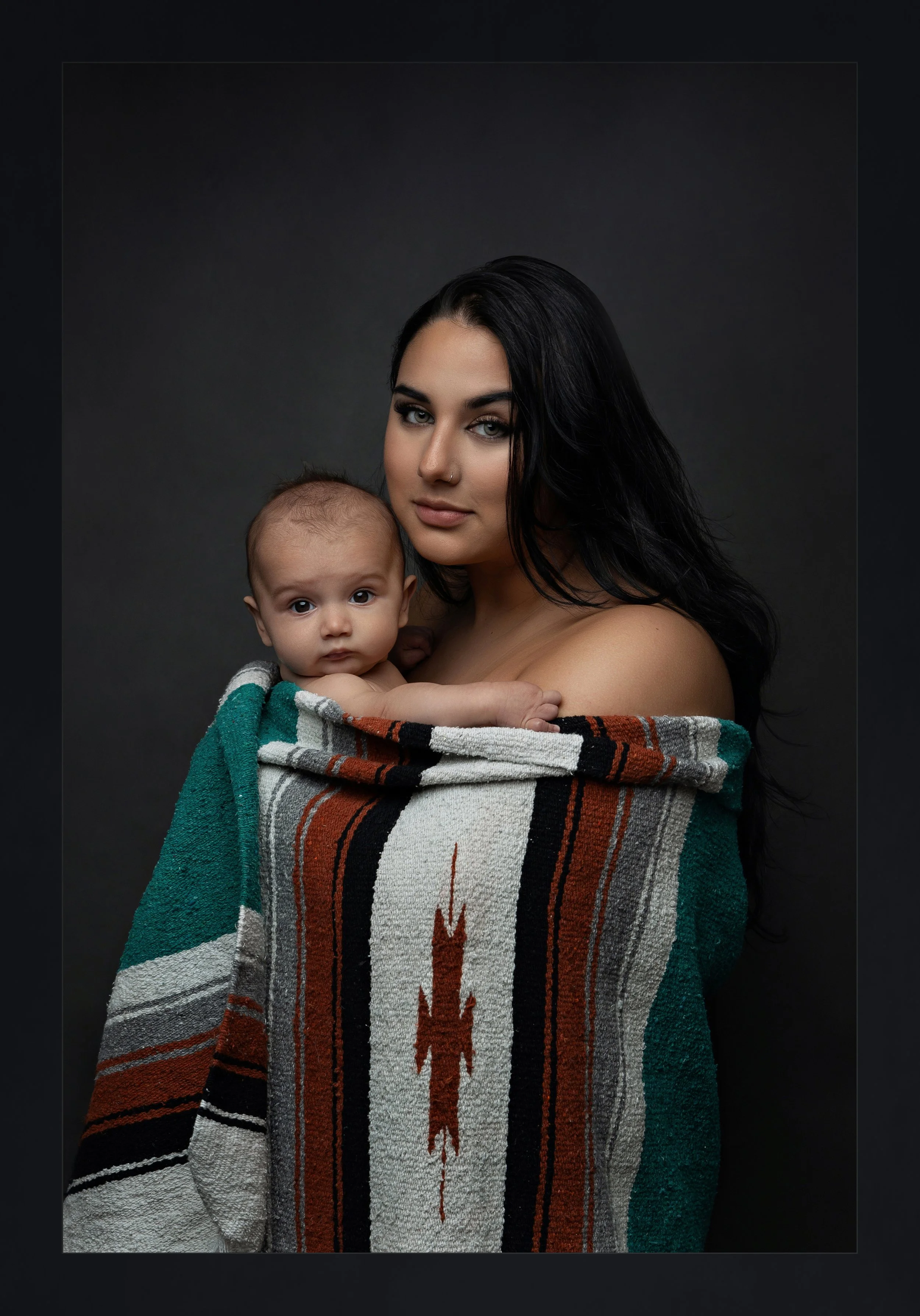Elegant studio portrait of a mother and child wrapped ina vibrant Southwestern-style blanket, showcasing a tender and intimate moment. The mother's captivating eyes adn serene expression paired with the child's innocent gaze create a powerful image o