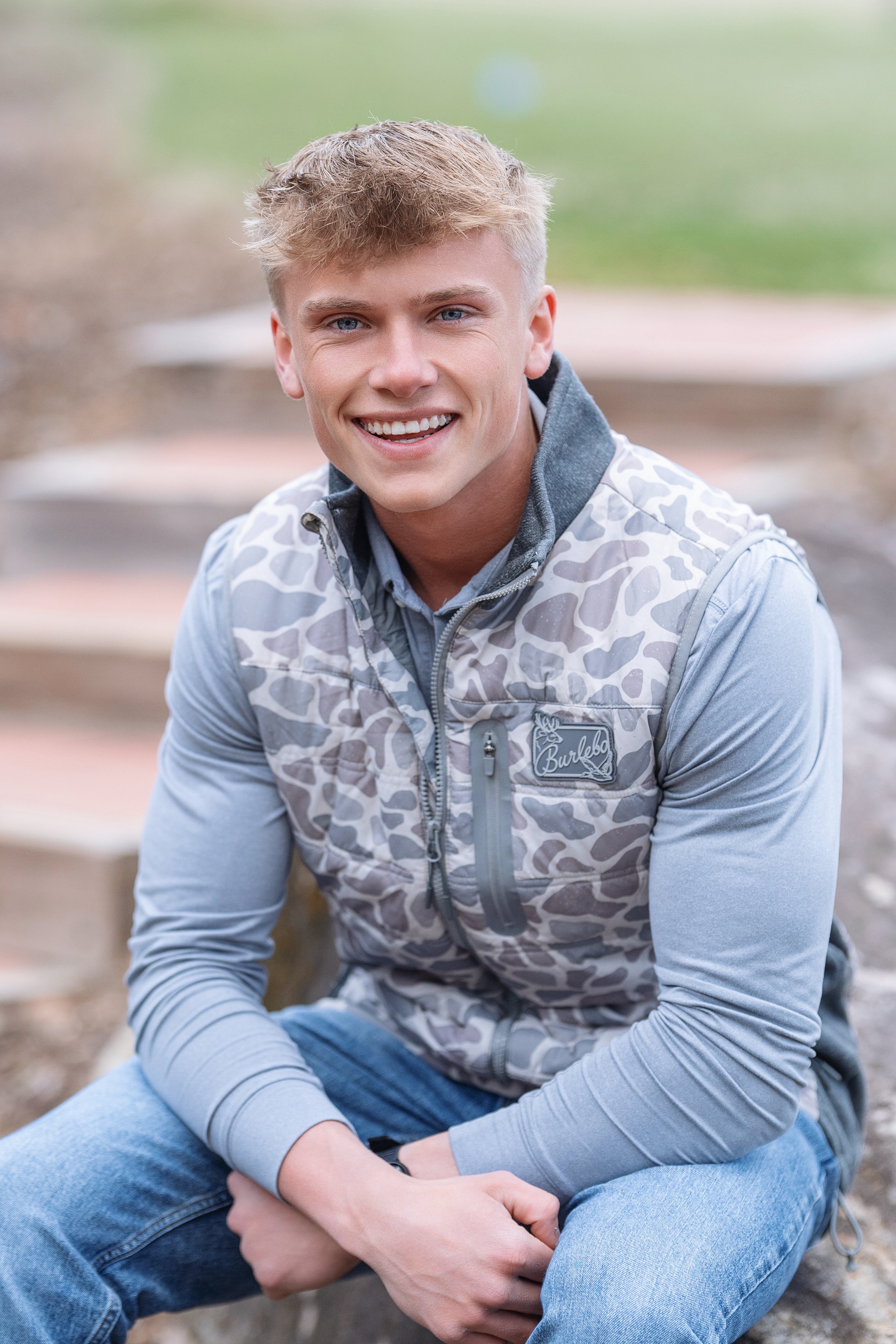 Warm and inviting portrait of a young man with a captivating smile, seated outdoors in North Myrtle Beach, donning a fashionable camouflage print vest by Burlesco. The casual setting and his approchable demeanor make this image ideal for High school 