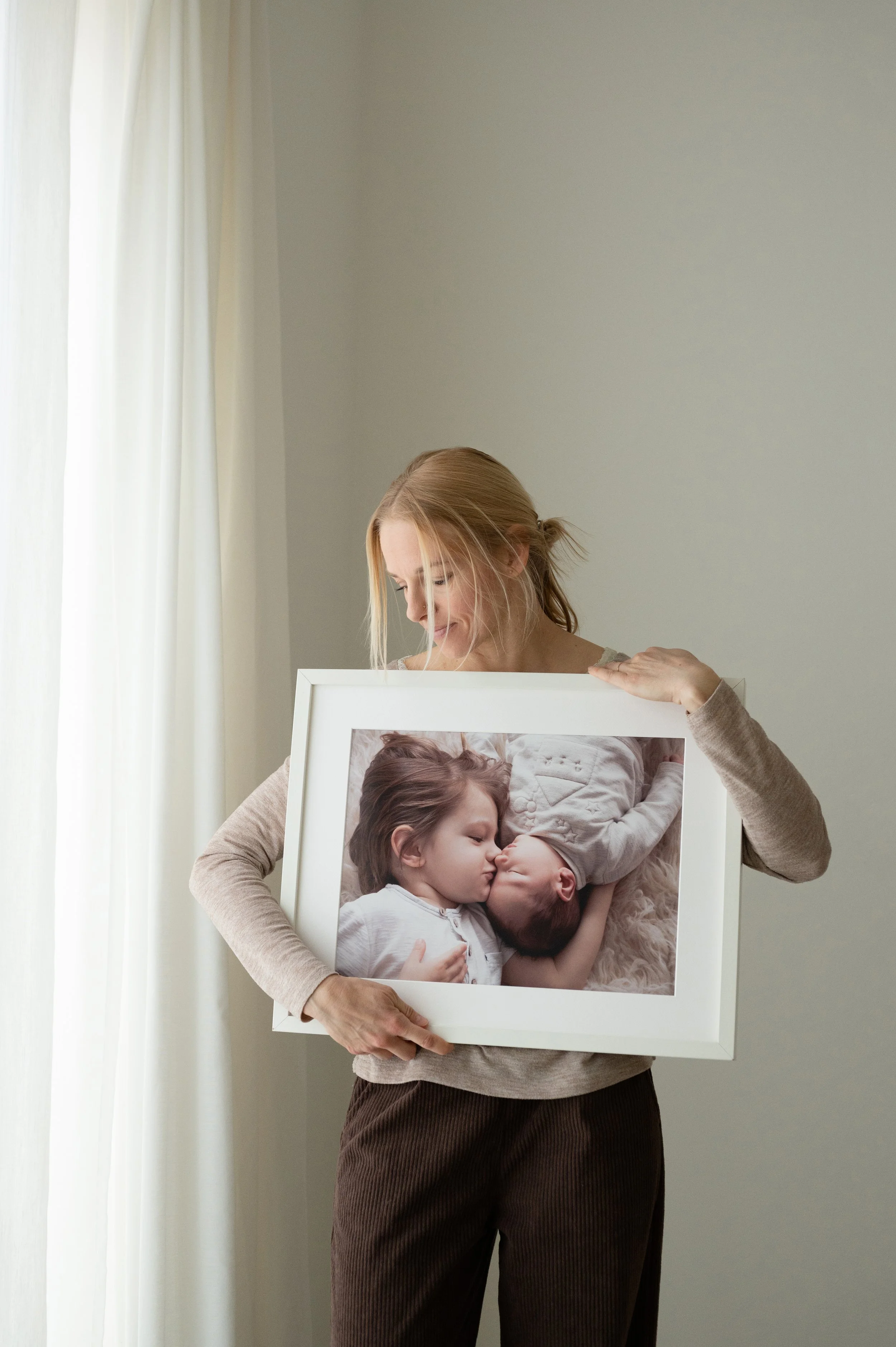 Una donna tiene in mano una cornice con una foto di due bambini che si baciano, uno bambino più grande e uno più piccolo, in un ambiente luminoso con tende bianche.