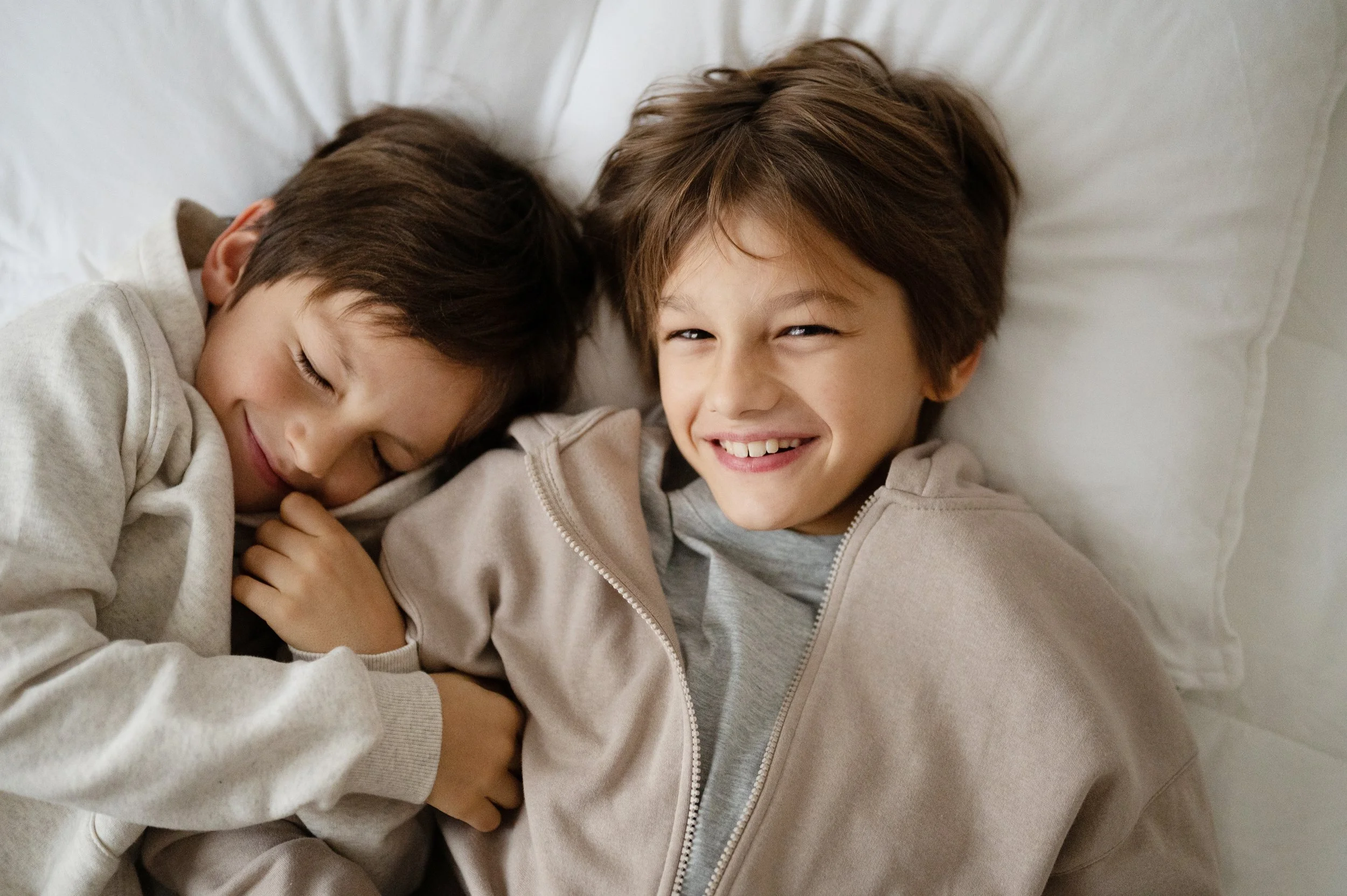 Due bambini con capelli castani e sorriso sono sdraiati sul letto, uno con gli occhi chiusi e l'altro con gli occhi aperti e un grande sorriso.