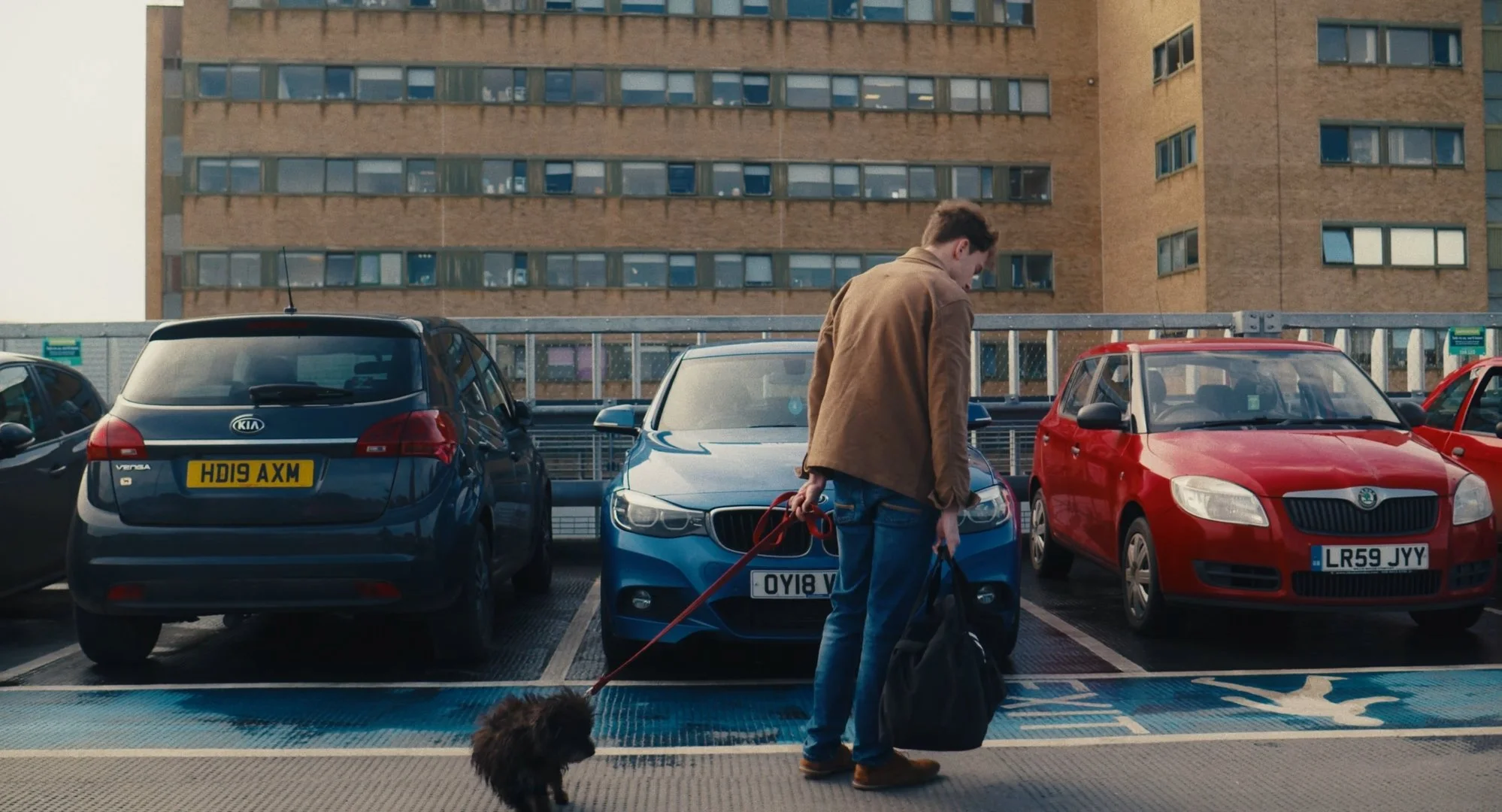 A person with brown hair, wearing a brown jacket and blue jeans, is standing in a parking lot, holding a leash attached to a dog. The parking lot has several parked cars, including a blue BMW and a red Skoda. In the background, there is a multi-story brick building with many windows.