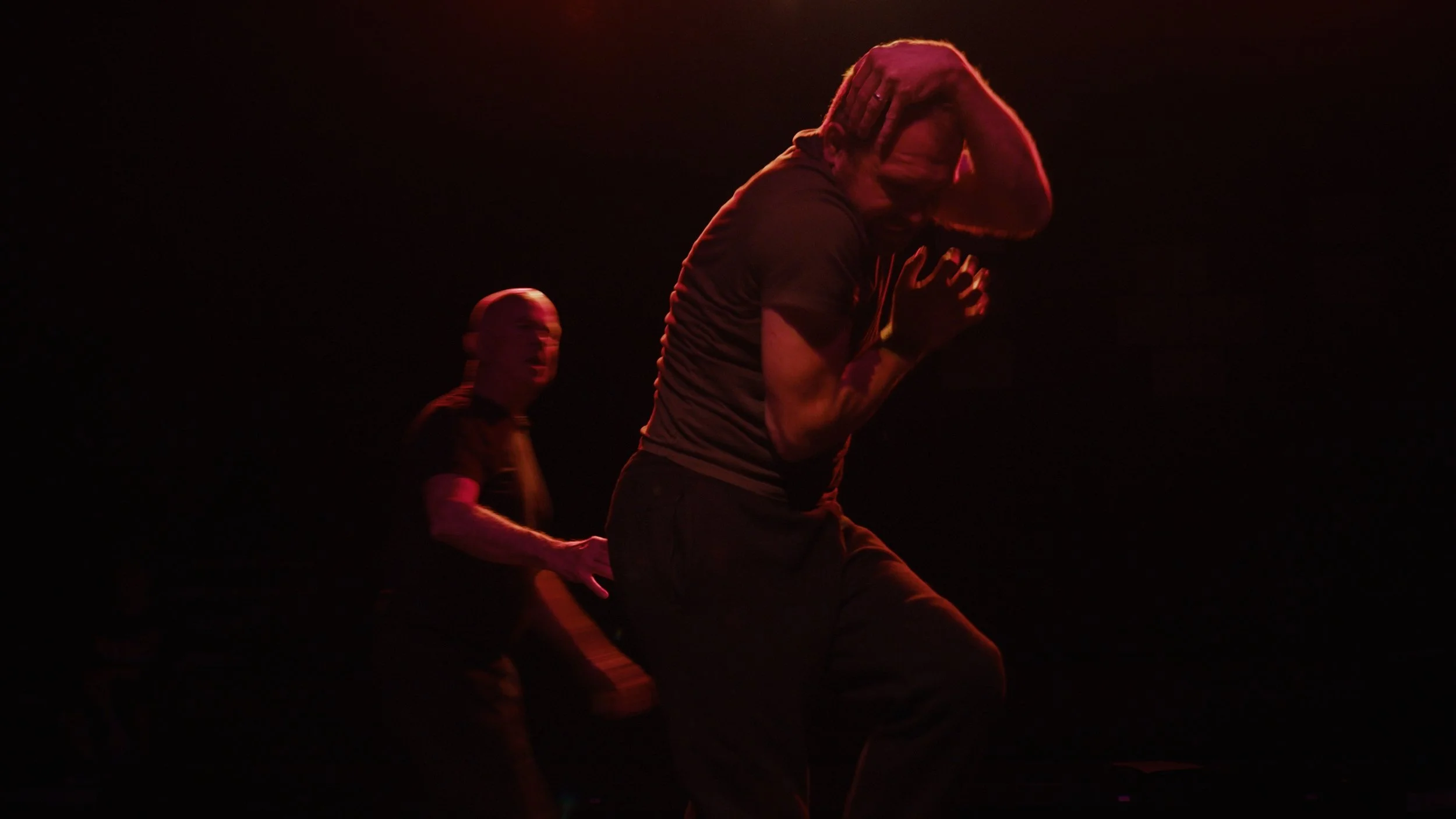 A man with long hair appears to be moving intensely with his fists clenched and head lowered, illuminated by red and orange lighting on stage with a dark background; a woman with short hair is in the background, slightly turned, wearing black, and also illuminated by stage lighting.