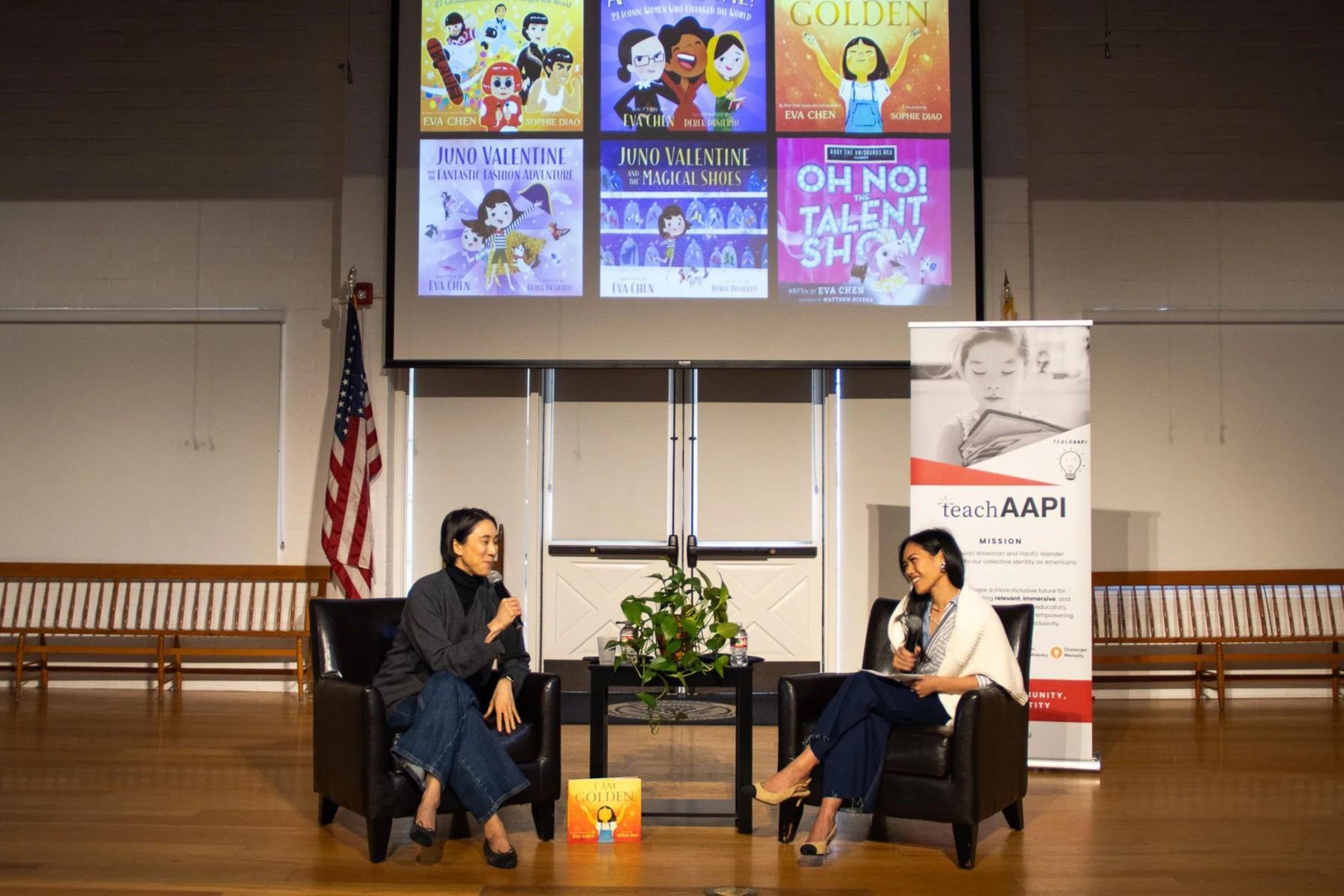 Two women sitting in black armchairs in a lecture hall, engaged in a conversation with microphones. Behind them, a large screen displays colorful book covers, and a banner with the logo and text 'teachAPI' is to the right. The American flag is to the