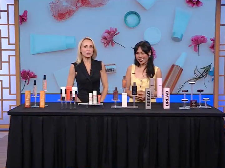 Two women standing behind a table with various skincare or cosmetic bottles, in front of a colorful background with flowers, skincare items, and beauty products displayed.