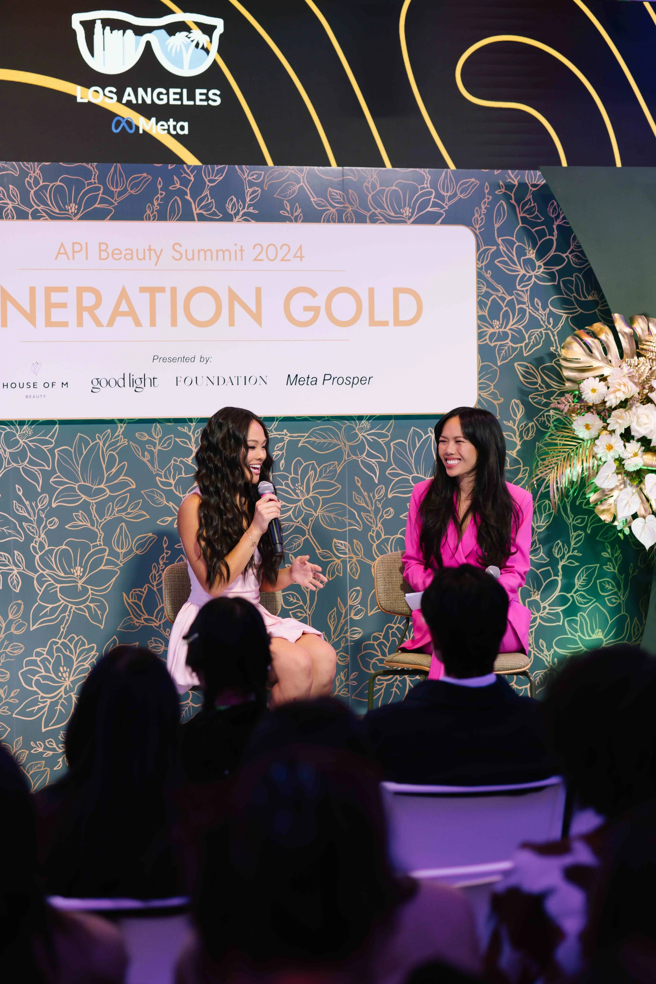 Two women seated on stage at the API Beauty Summit 2024, smiling and engaging in conversation. One holds a microphone, with the backdrop displaying a large sign reading 'Generation Gold' and floral design elements, and a screen above showing logos fo