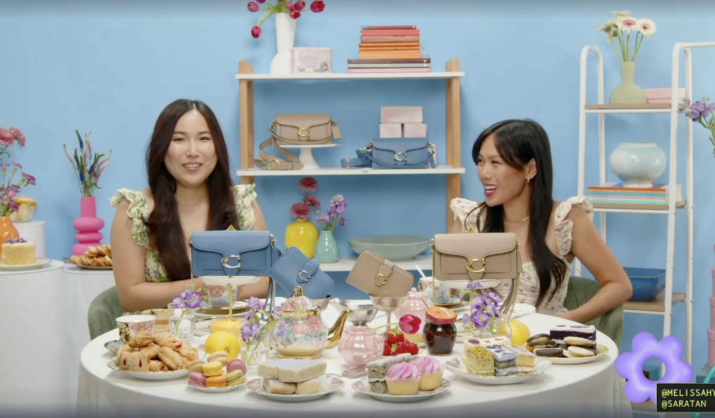 Two women sit at a table with various desserts, colorful handbags, and flowers, in front of a light blue wall with shelves holding more handbags and decorative items.