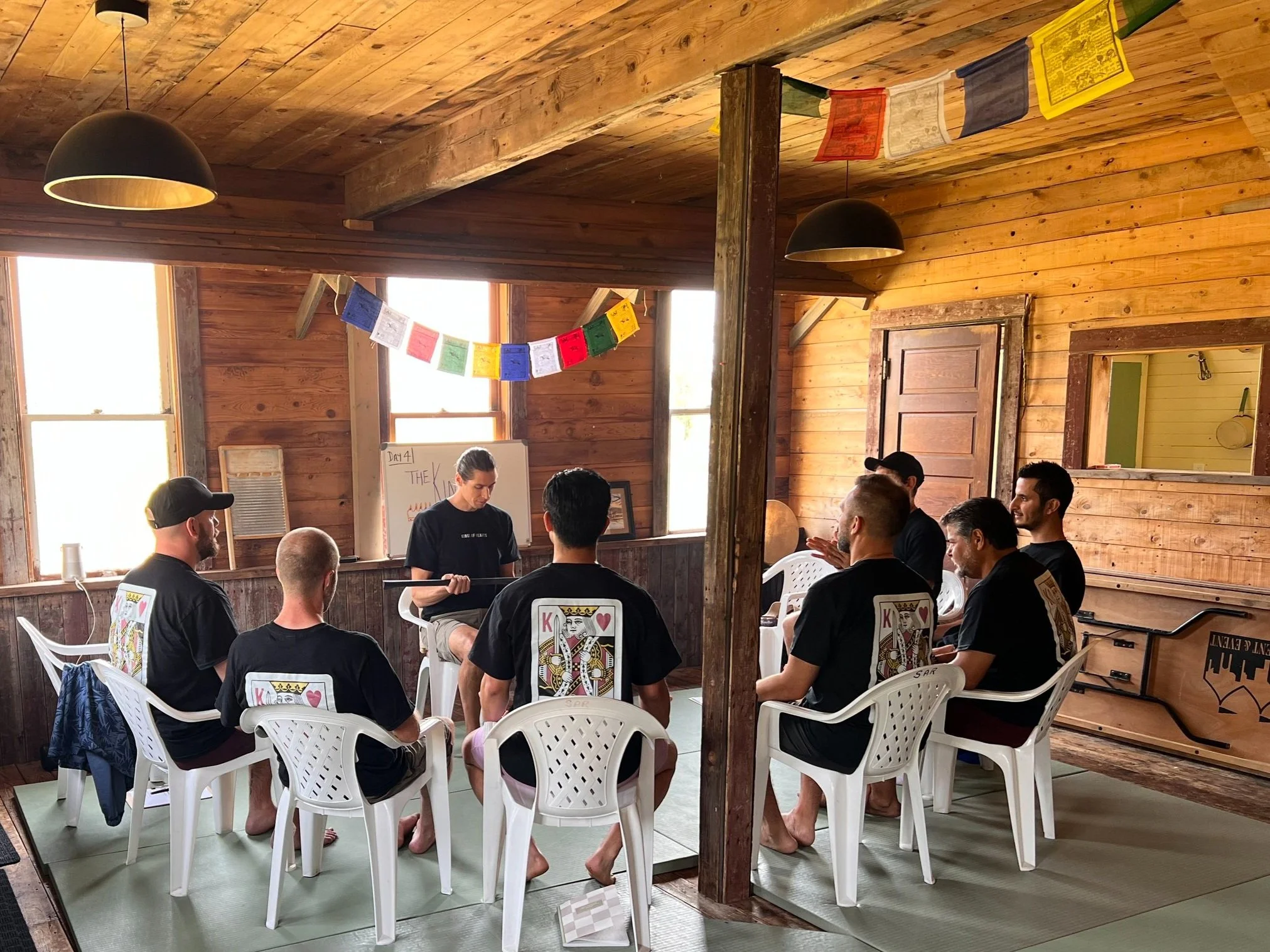 A group of people in a wooden room sitting in a circle on white plastic chairs. They are wearing matching black shirts with a playing card design on the back. A person is at the front near a whiteboard, likely leading a discussion or class. Colorful flags hang from the ceiling.