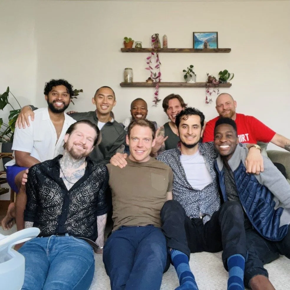 Group of nine men sitting on the floor in a living room, smiling, with plants and decor on shelves in the background.