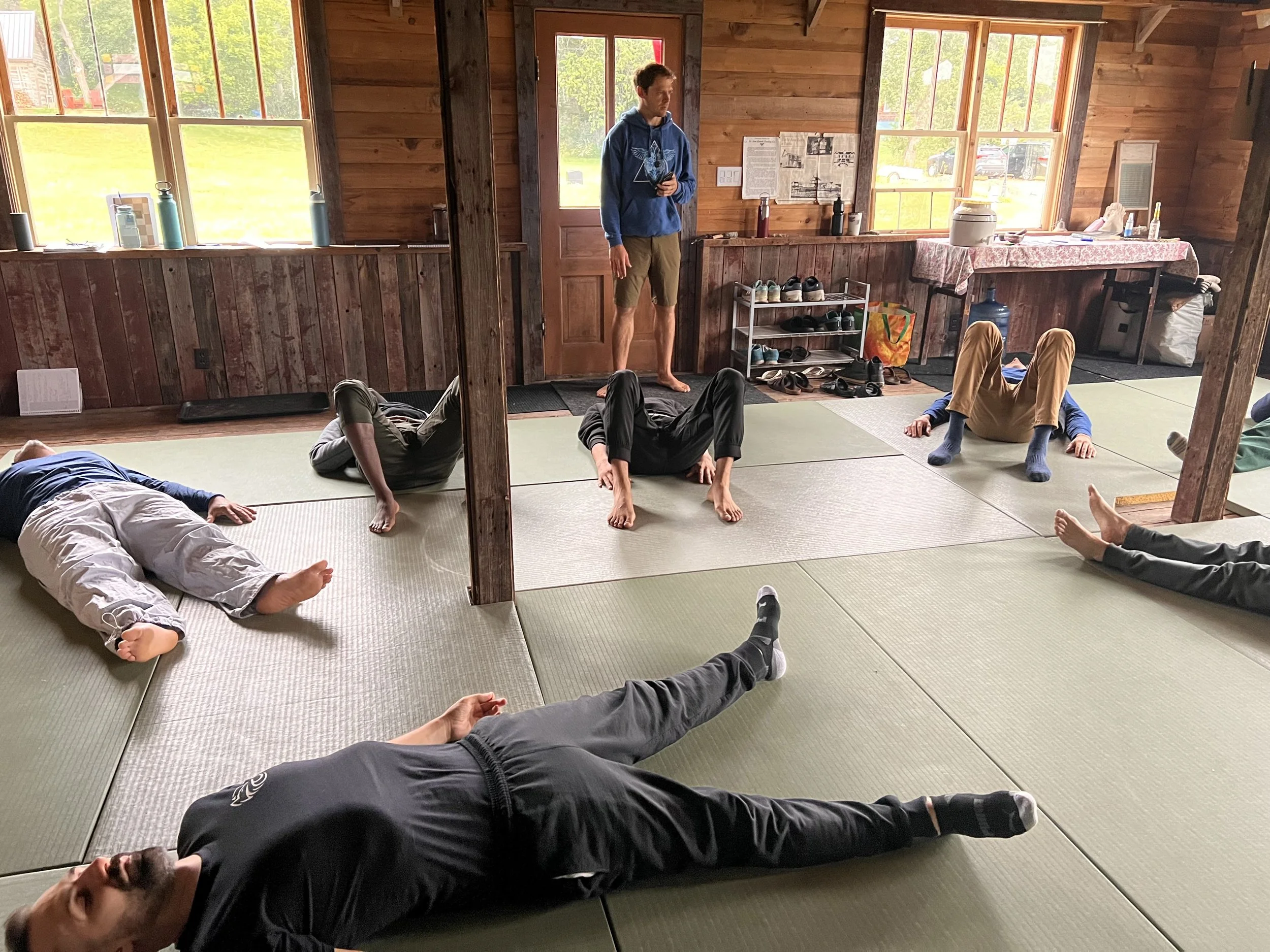 A group of people lying on mats in a rustic wooden room, practicing a relaxation exercise or meditation. A person is standing and instructing the session. The room has large windows and various items on a table in the background.