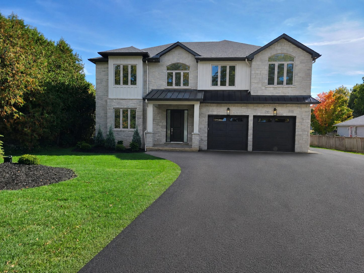 A modern two-story house with a stone exterior, black garage doors, and a black front door. The house has a gray shingle roof, large windows, and a manicured lawn with some small shrubs. There is a paved driveway leading to the garage and a small garden bed with mulch and plants on the left side.