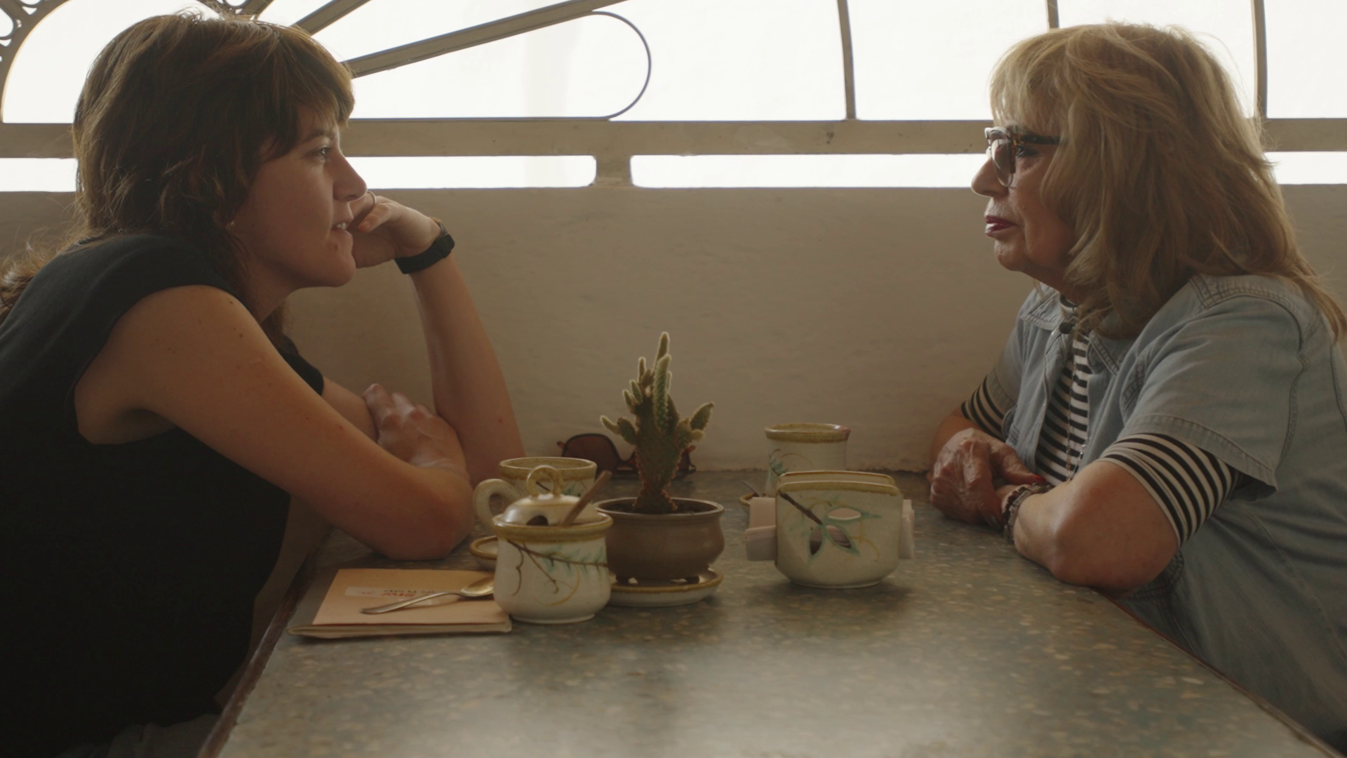 Two women sitting across from each other at a lunch table, engaging in conversation. The table has a small potted cactus, mugs, and a menu. Window with blinds is behind.