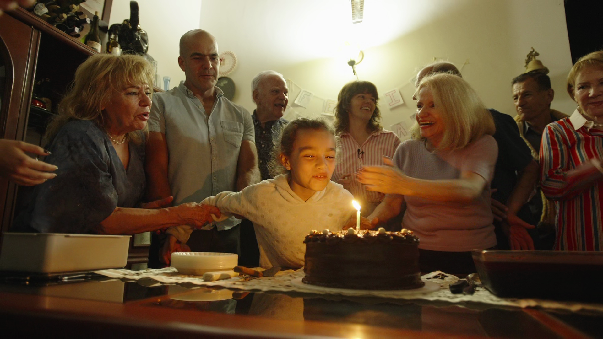 A group of people celebrating a birthday around a table with a chocolate cake with lit candles. The girl in the center is blowing out the candle, surrounded by smiling adults.