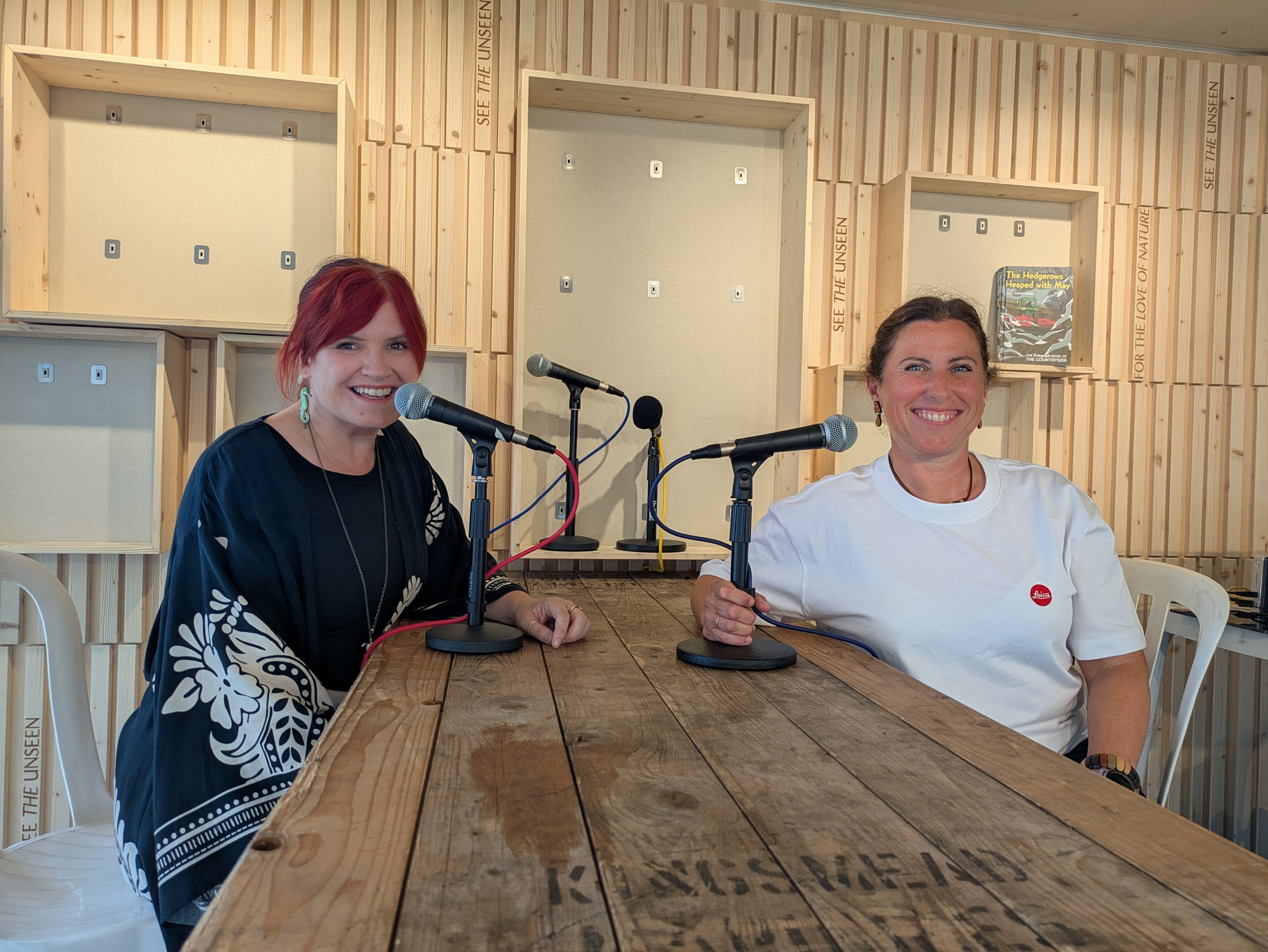 Kelly Keegan smiling into the camera, sat opposite Penny Green who is also smiling at the camera. Both women have microphone in front of them and are recording live from the podcast stage at Global Birdfair