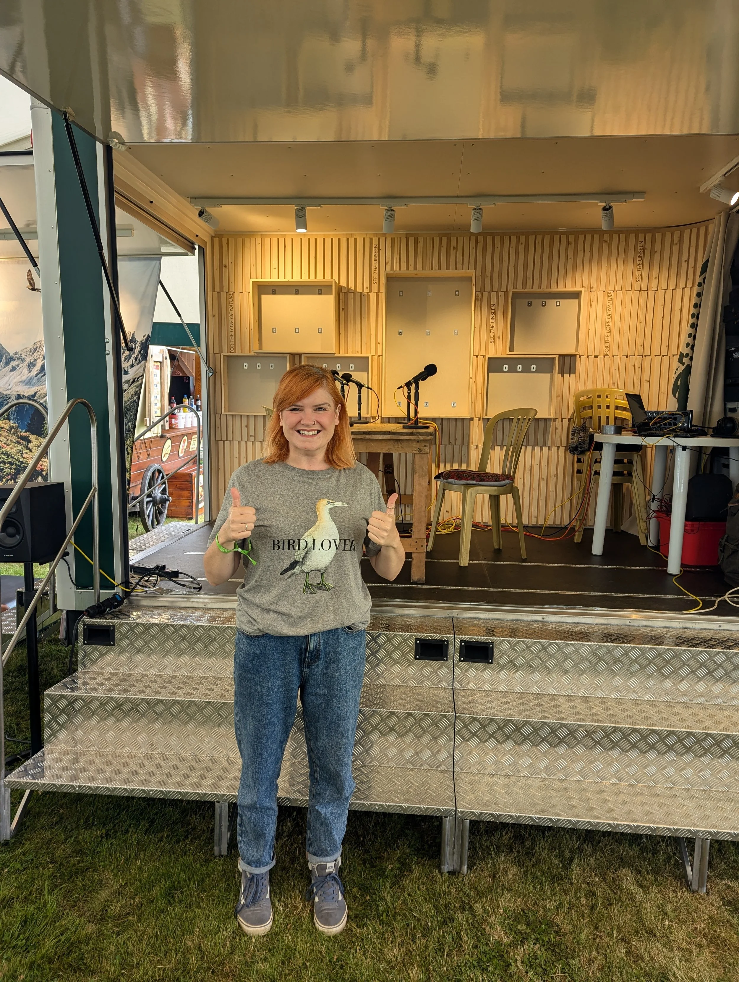Public speaker and podcast host Kelly Keegan at a live event discussing nature, storytelling and community engagement, holding two thumbs up and grinning in front of the stage.