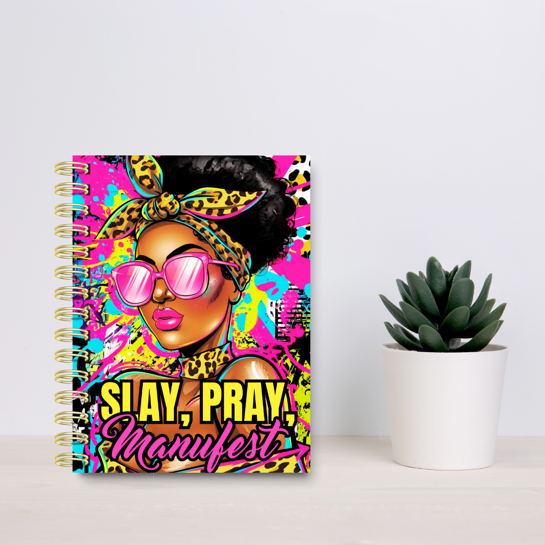 Colorful notebook with a pop art style illustration of a woman wearing leopard print accessories and pink sunglasses, with the text 'SAY, PRAY, MANIFEST' on the cover, placed next to a potted succulent plant on a white surface.