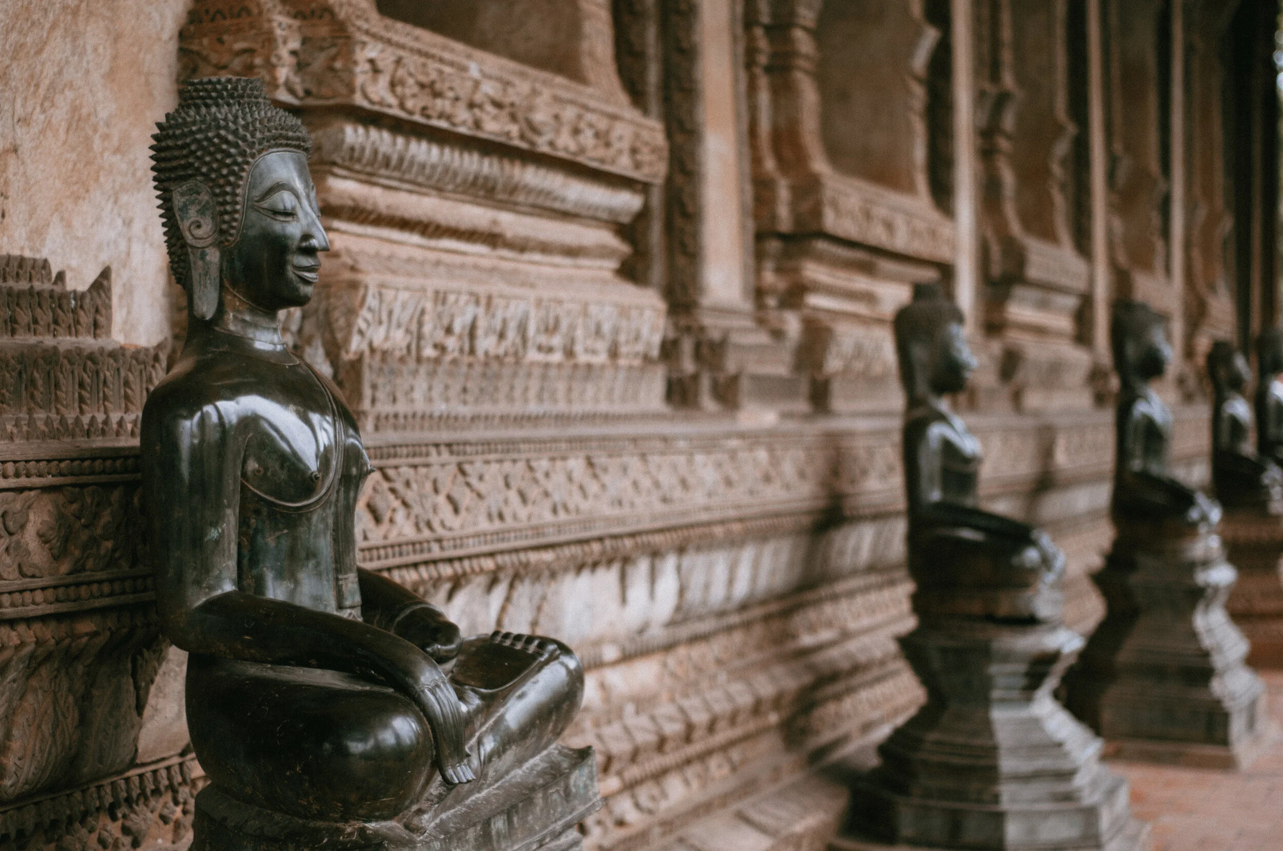 Four dark-colored seated Buddha statues with intricate carvings behind each, made of wood or metal, arranged in a row inside a temple with carved wooden walls.
