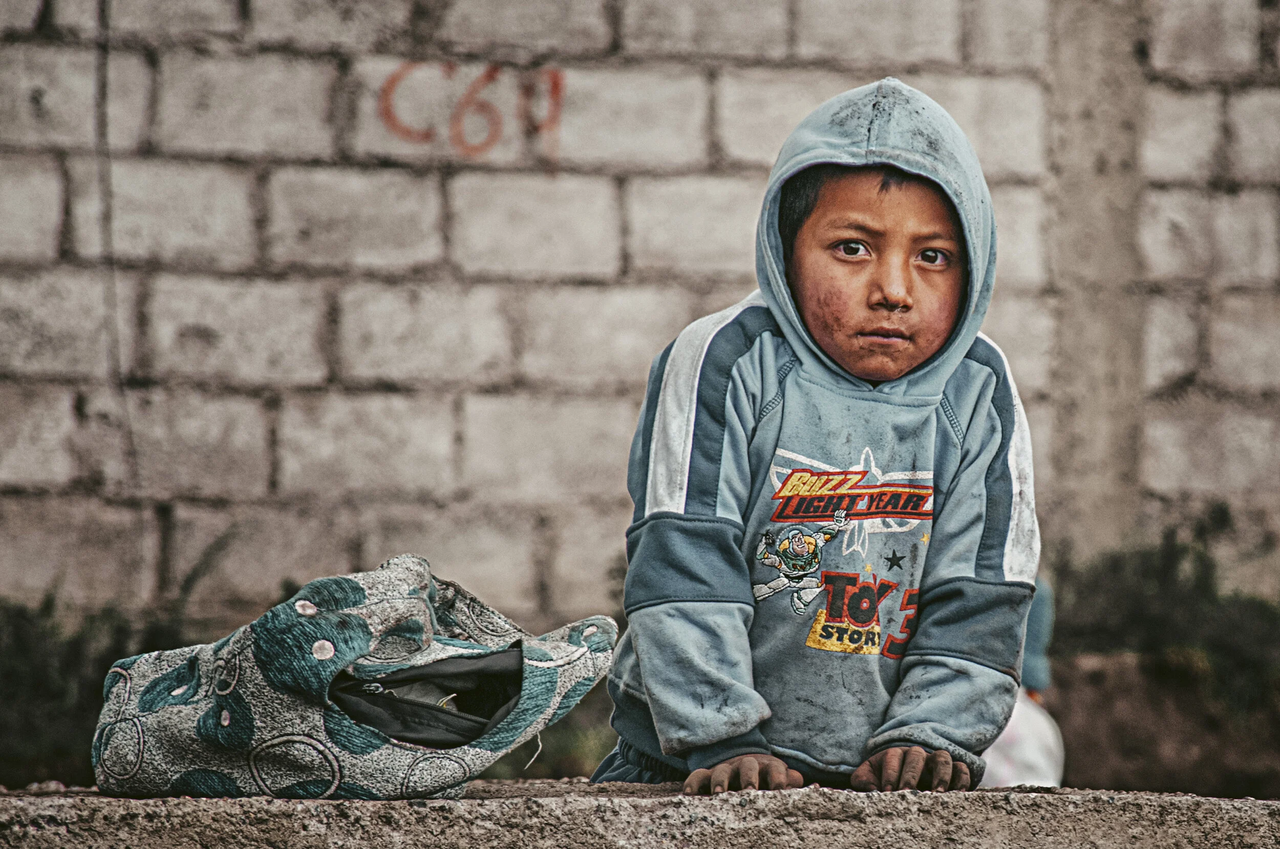 Young boy with dirty face and clothes, wearing a hooded sweatshirt with a cartoon design, leaning on a concrete ledge next to a patterned bag, with a brick wall in the background.
