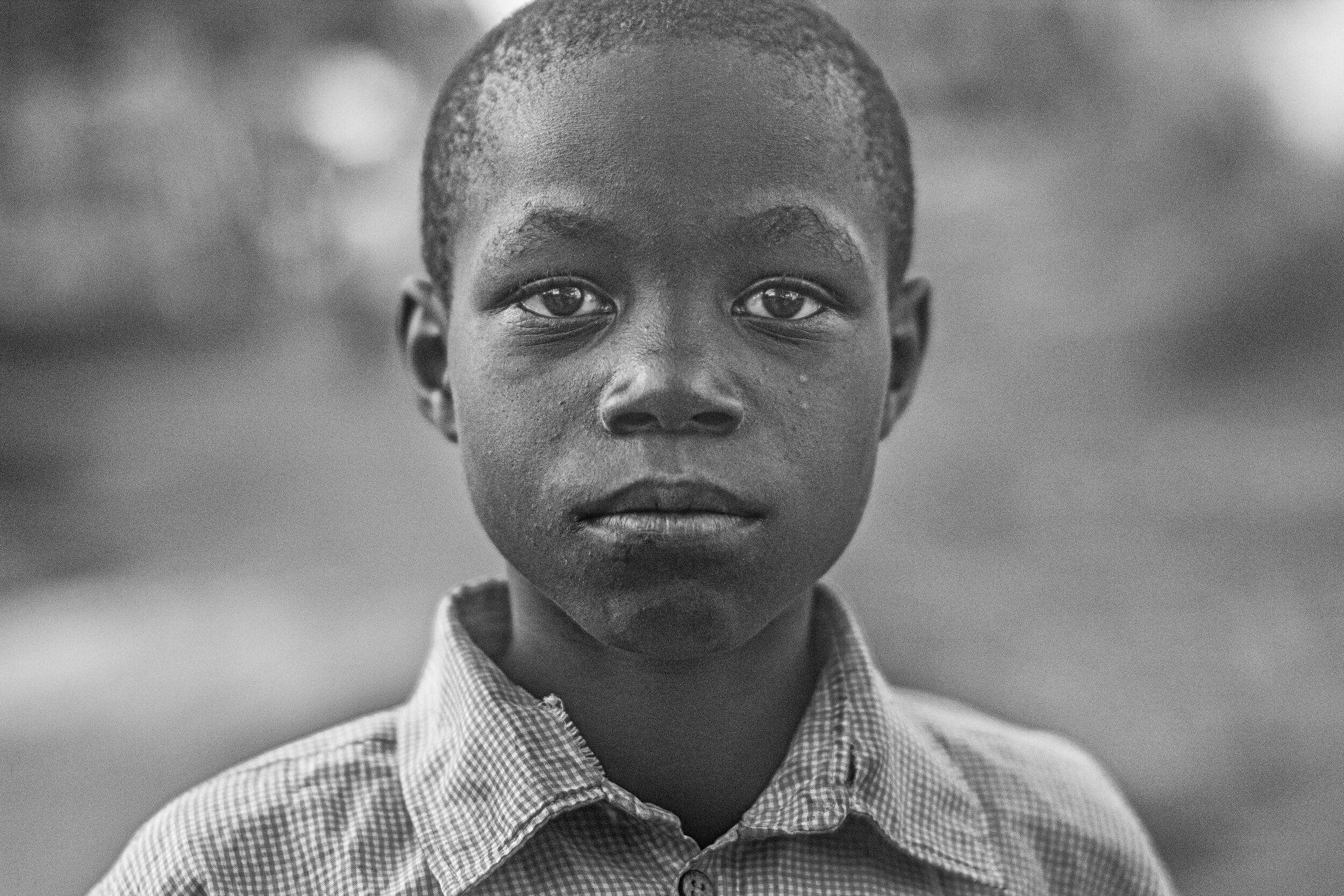 A black-and-white portrait of a young boy with short hair, wearing a checkered shirt, looking directly into the camera.